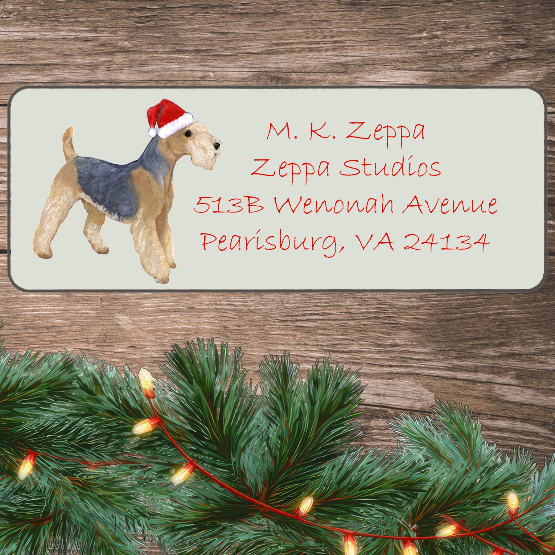 An illustration of a Lakeland Terrier wearing a Santa Hat on an ivory return address label set off by a wood surface with holiday greenery background.