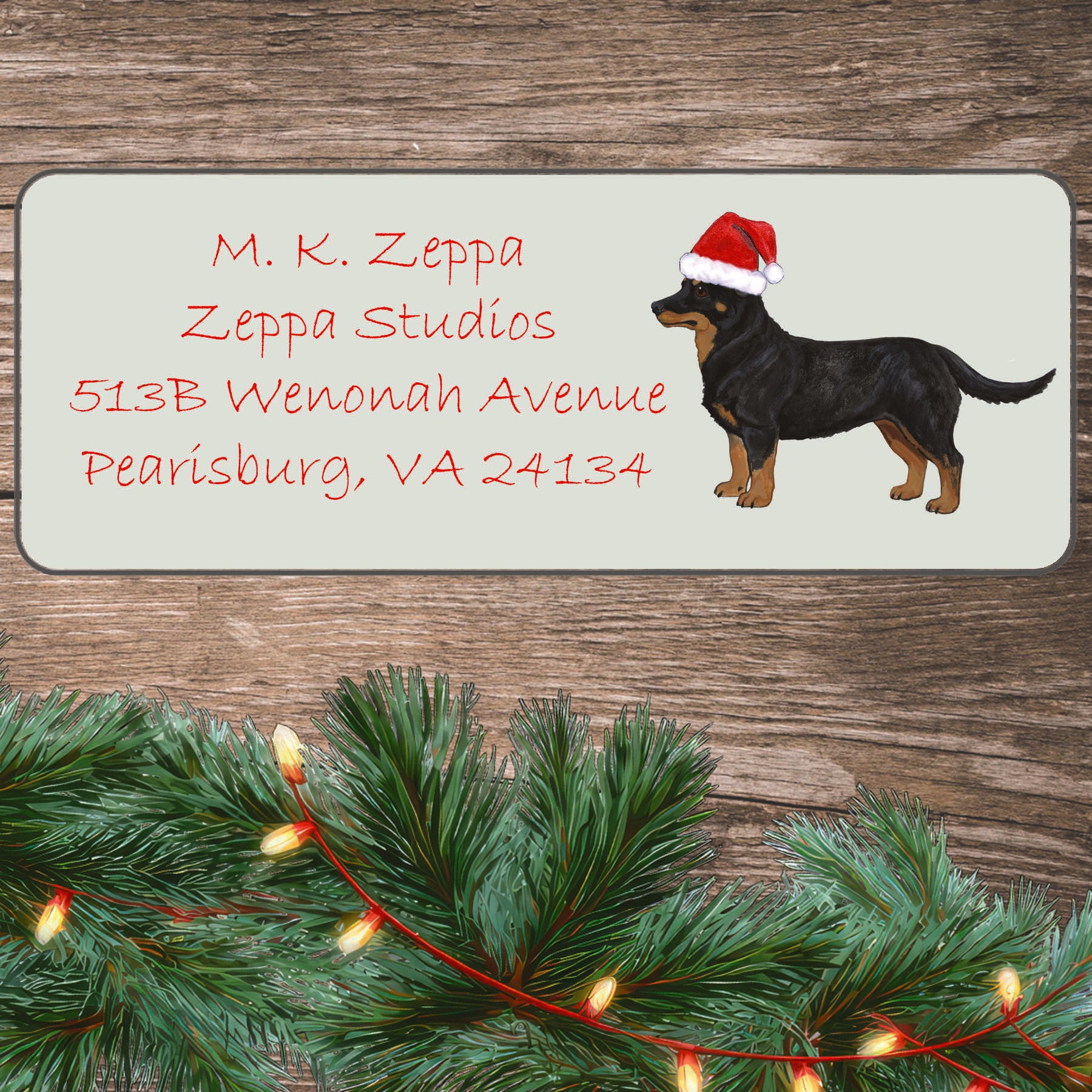 An illustration of a Lancashire Heeler wearing a Santa Hat on an ivory return address label set off by a wood surface with holiday greenery background.