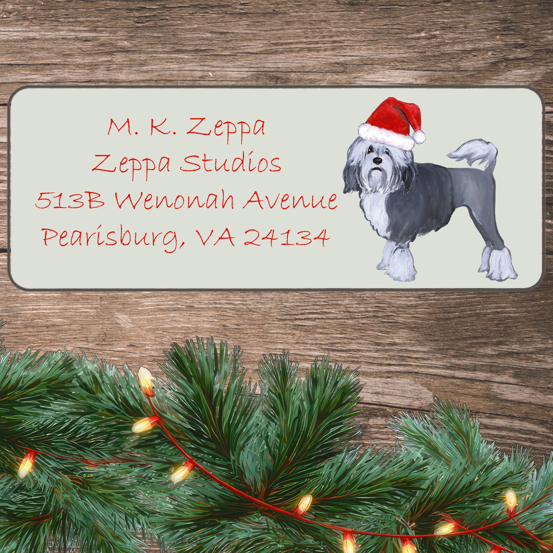 An illustration of a Lowchen wearing a Santa Hat on an ivory return address label set off by a wood surface with holiday greenery background.