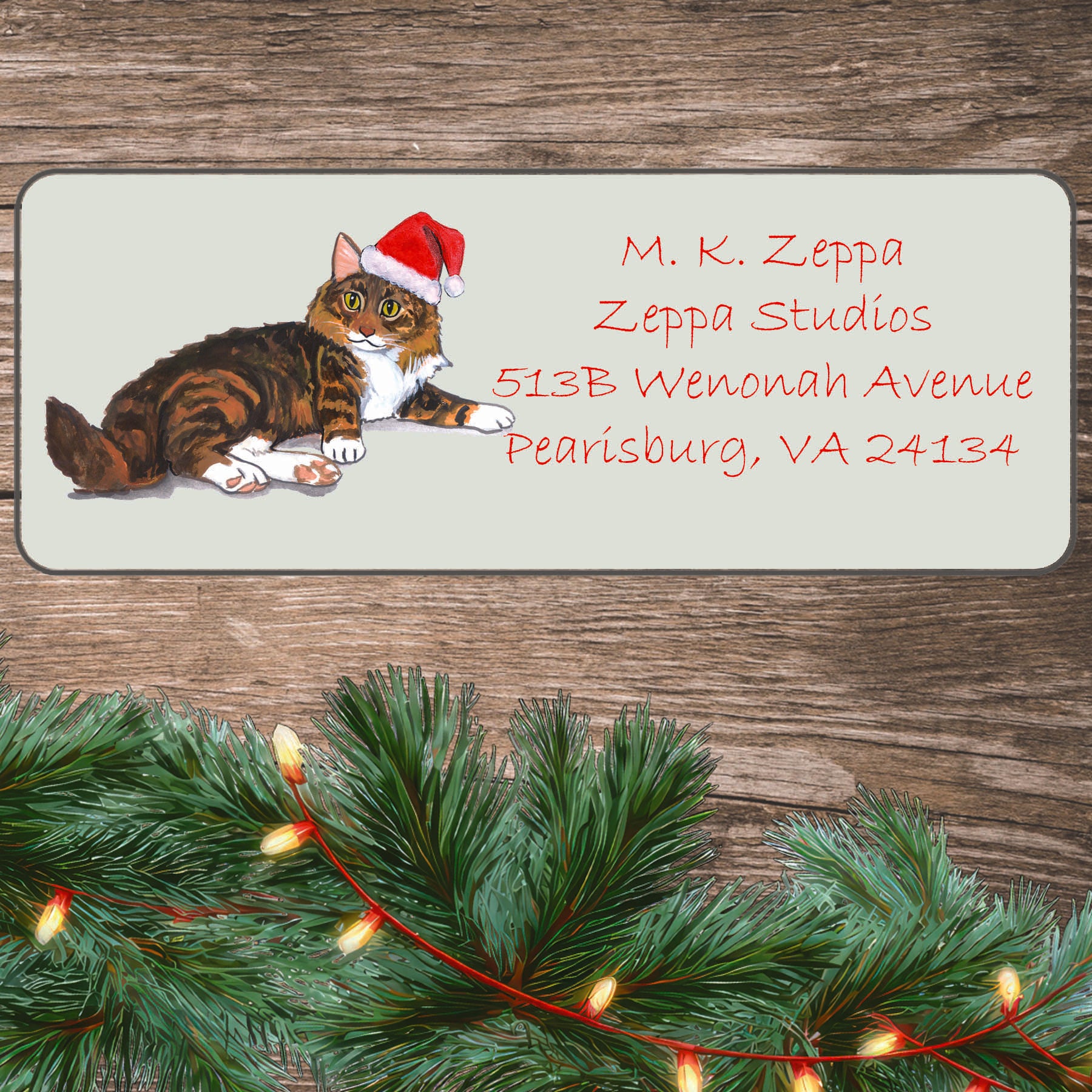 An illustration of a Maine Coon Cat wearing a Santa Hat on an ivory return address label set off by a wood surface with holiday greenery background.