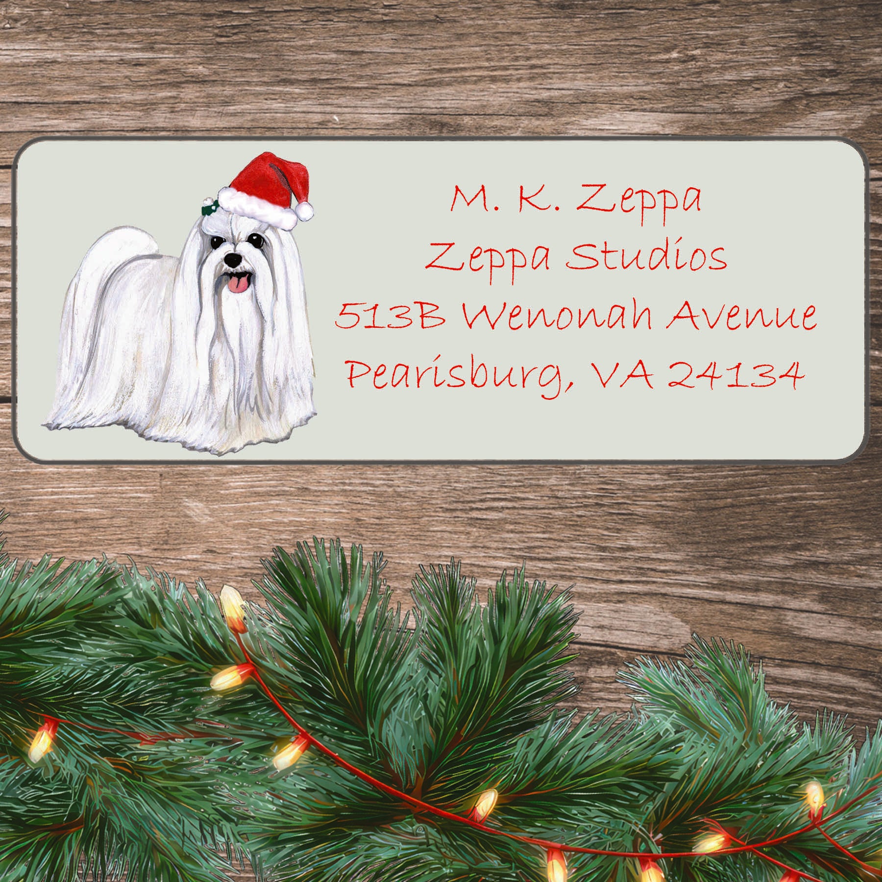 An illustration of a Maltese wearing a Santa Hat on an ivory return address label set off by a wood surface with holiday greenery background.
