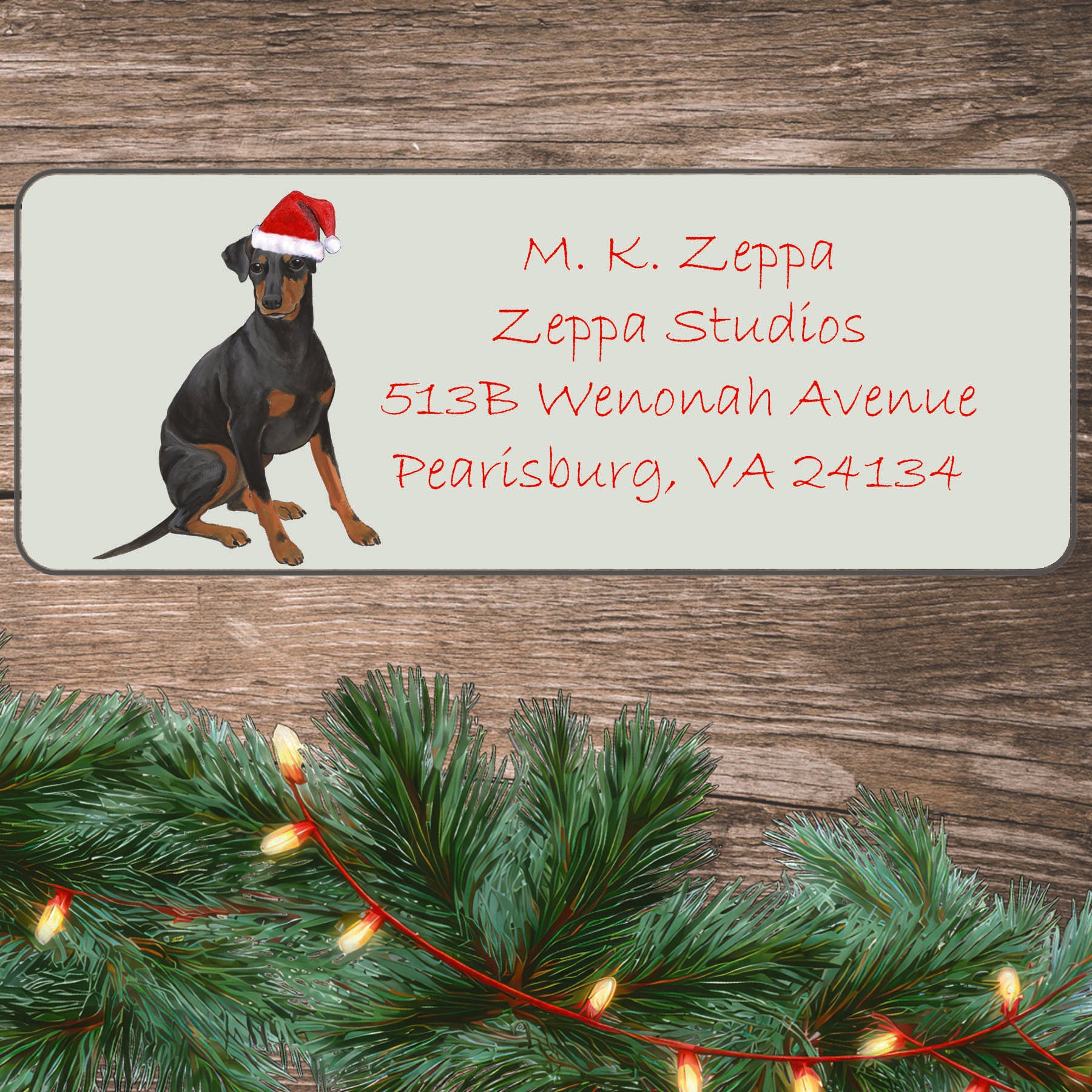 An illustration of a Manchester Terrier wearing a Santa Hat on an ivory return address label set off by a wood surface with holiday greenery background.