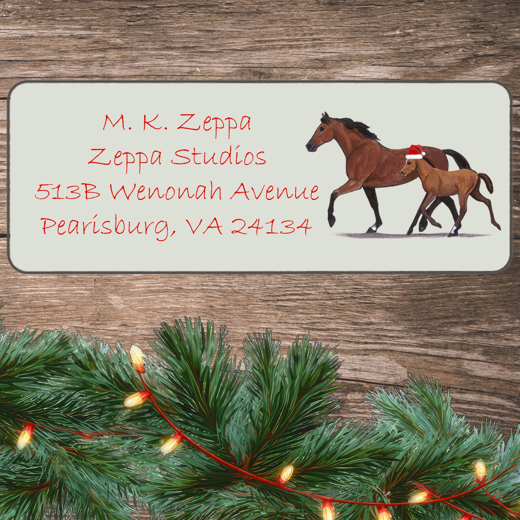 An illustration of a mare with a foal wearing a Santa Hat on an ivory return address label set off by a wood surface with holiday greenery background.