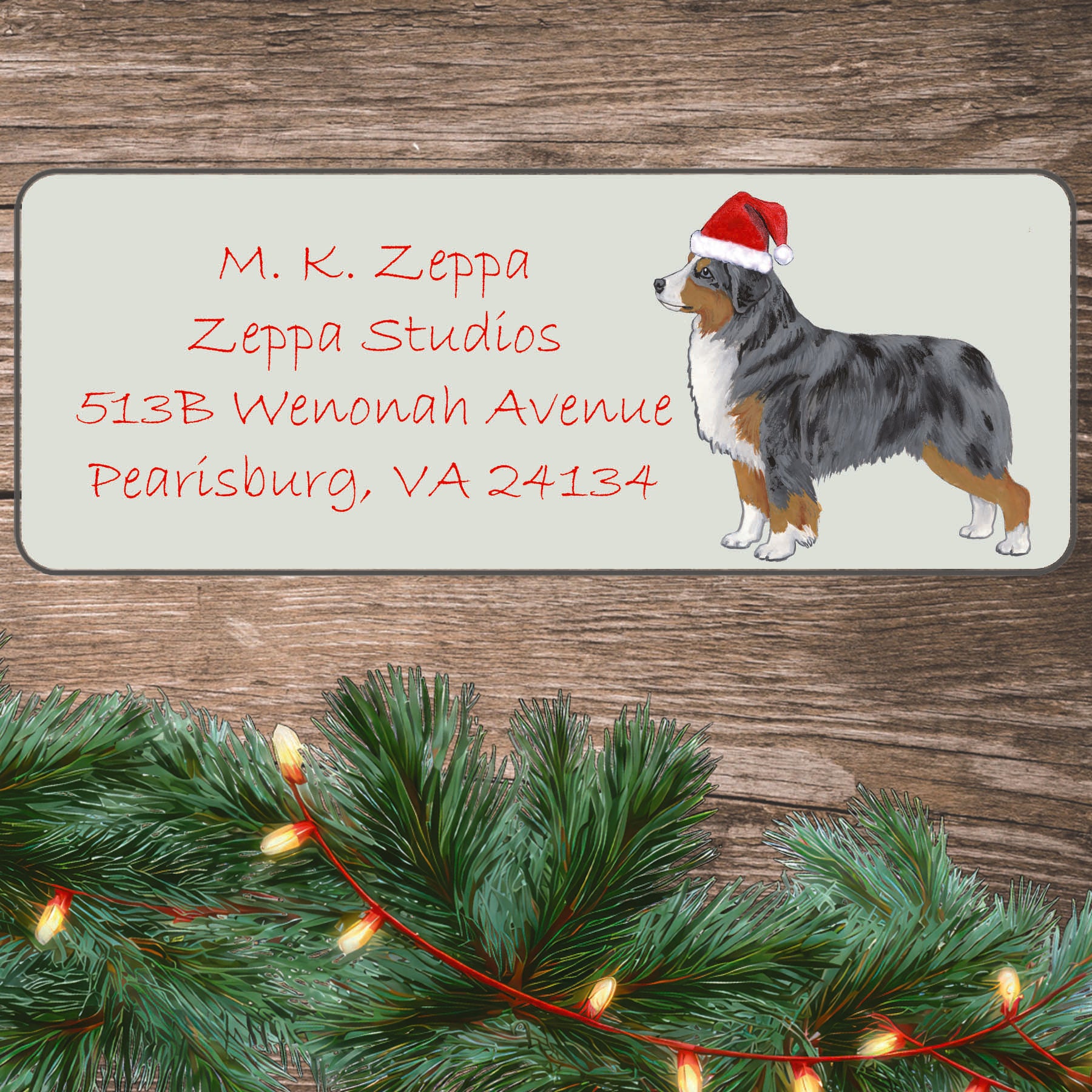 A Miniature American Shepherd in a Santa Hat illustration on an ivory return address label set off by a wood surface with holiday greenery background.