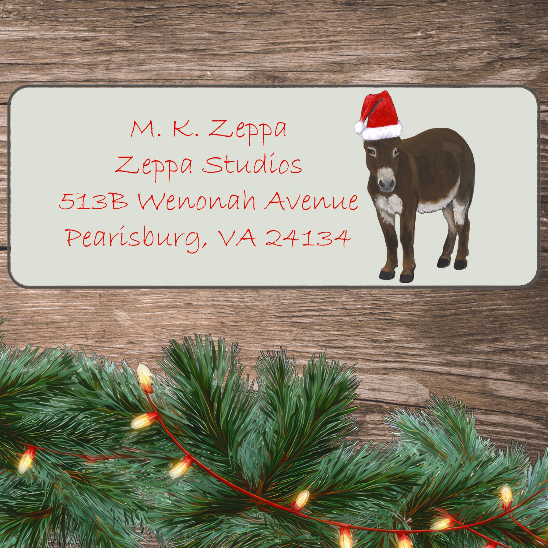 An illustration of a Miniature Donkey wearing a Santa Hat on an ivory return address label set off by a wood surface with holiday greenery background.