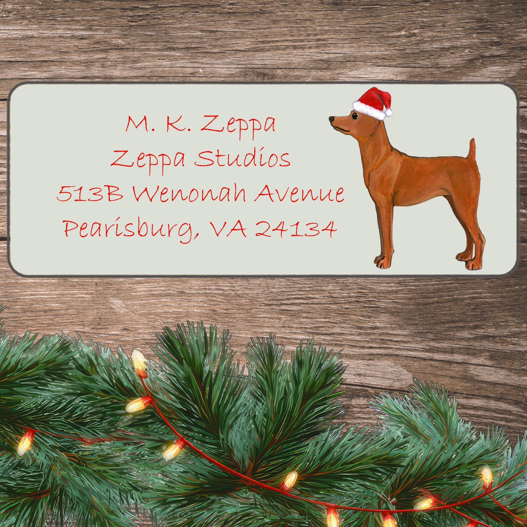 An illustration of a red Miniature Pinscher wearing a Santa Hat on an ivory return address label set off by a wood surface with holiday greenery background.
