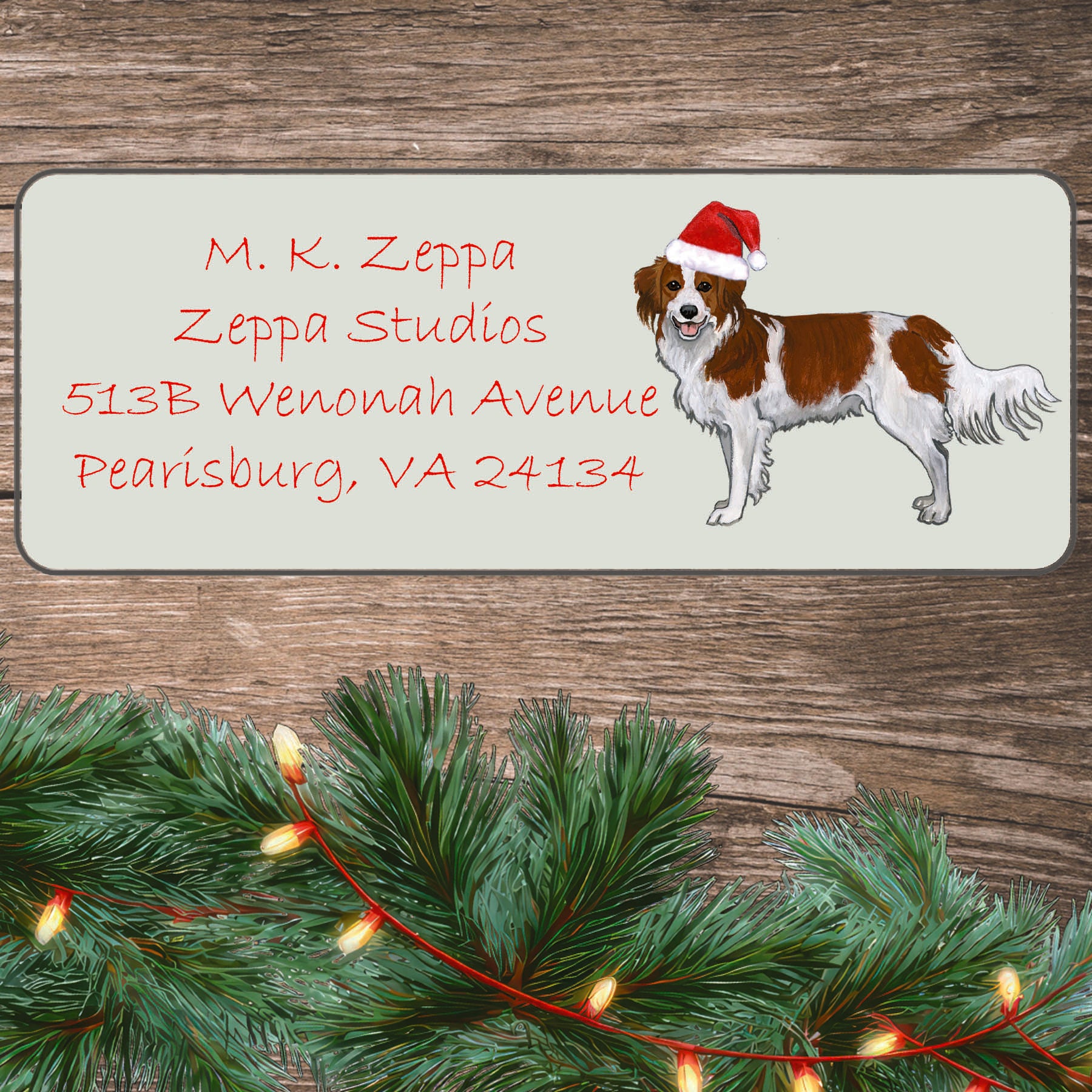 An illustration of a Nederlandse Kooikerhondje wearing a Santa Hat on an ivory return address label set off by a wood surface with holiday greenery background.