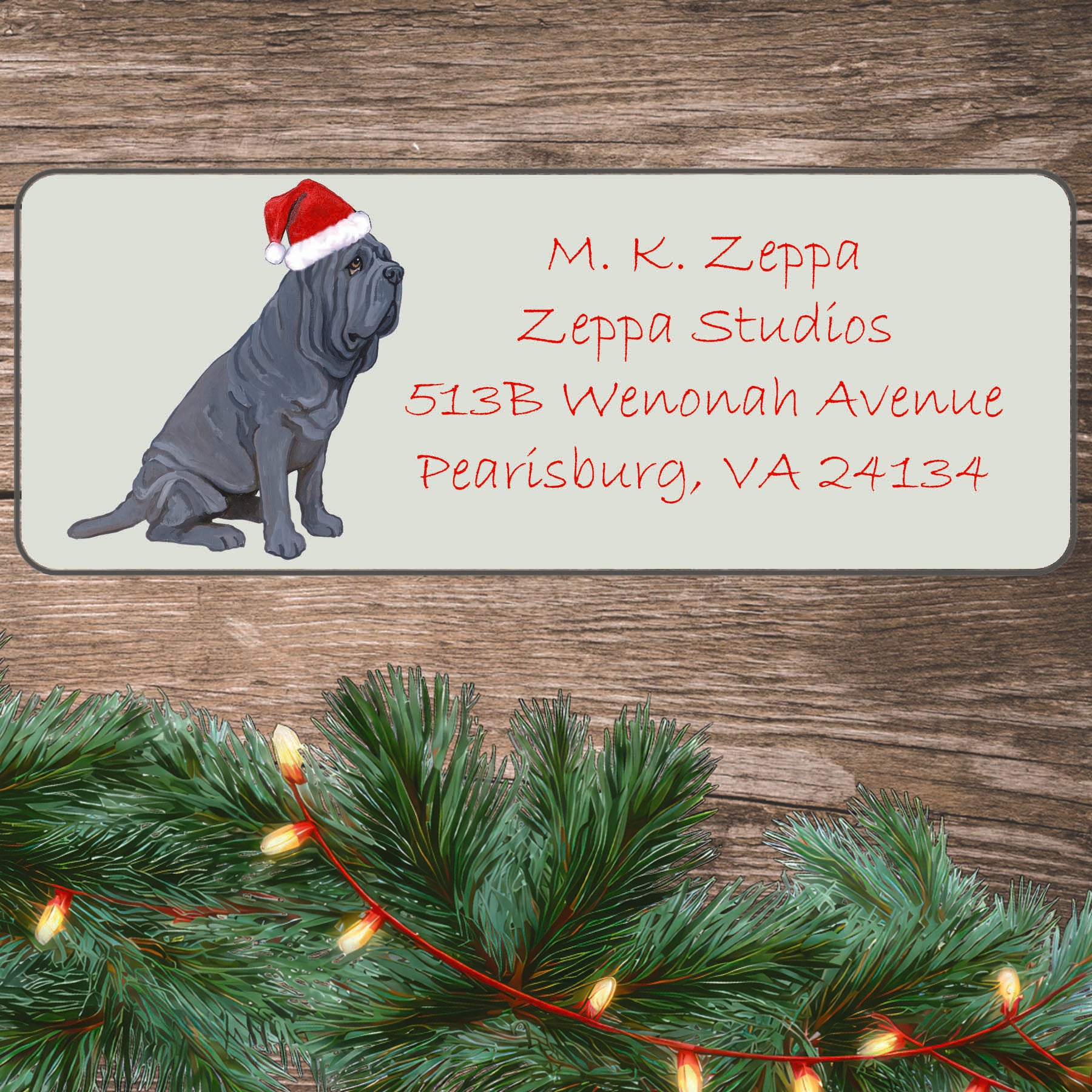 An illustration of a Neopolitan Mastiff wearing a Santa Hat on an ivory return address label set off by a wood surface with holiday greenery background.