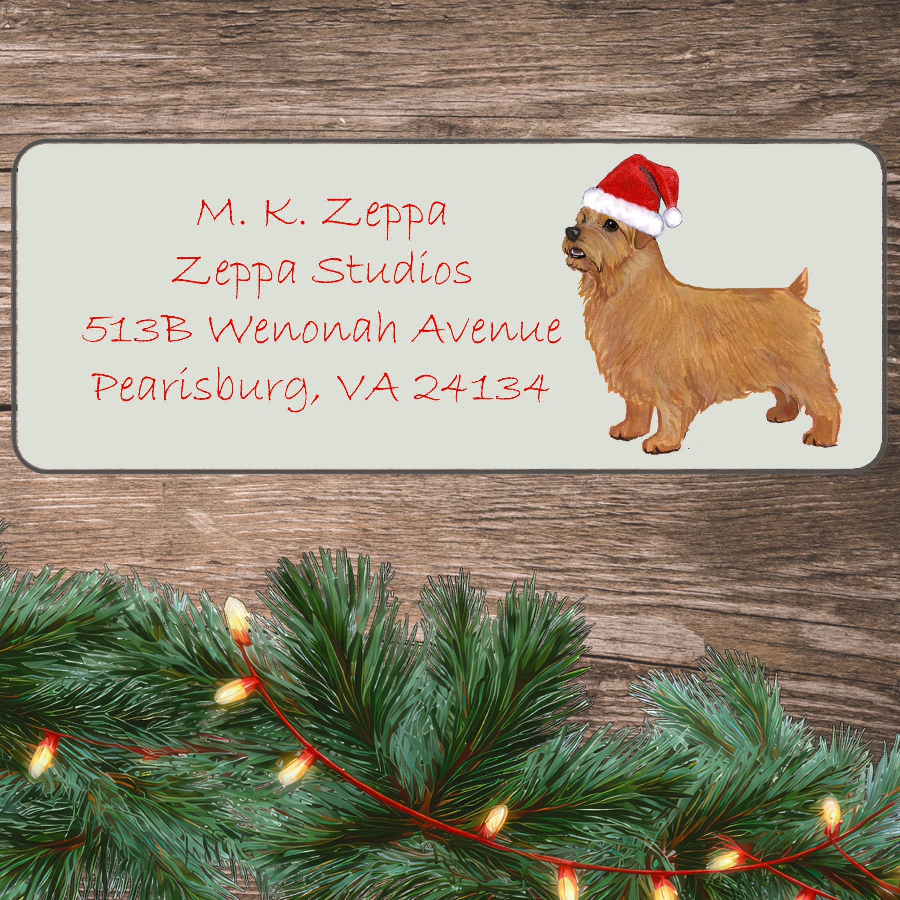 An illustration of a Norfolk Terrier wearing a Santa Hat on an ivory return address label set off by a wood surface with holiday greenery background.