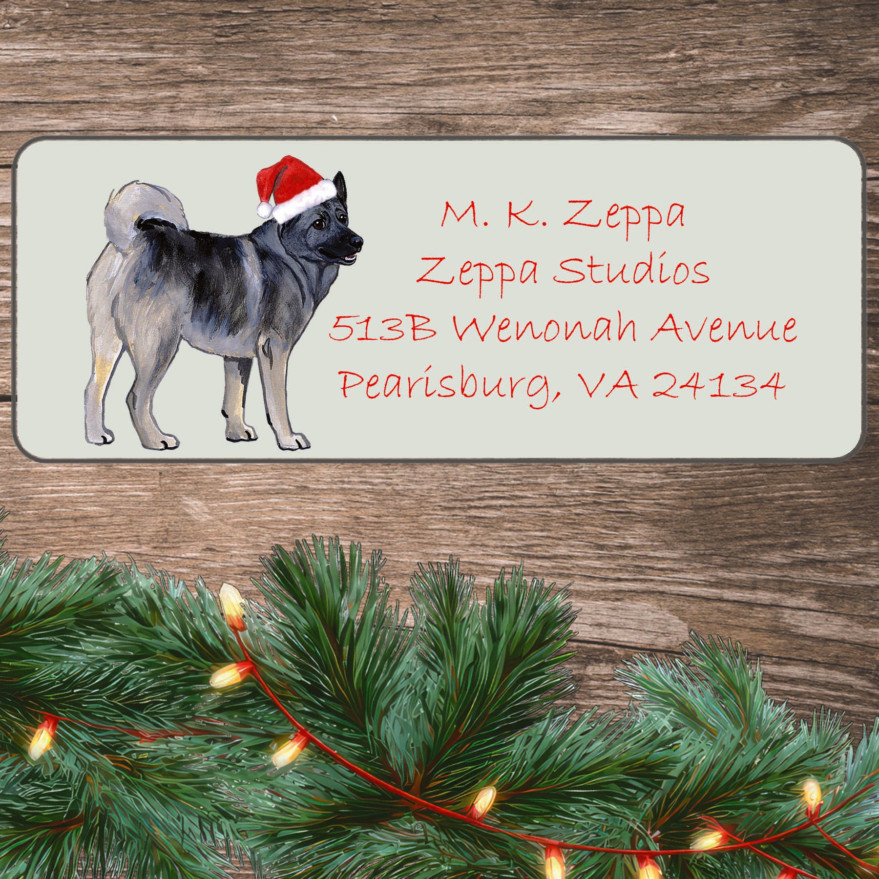 An illustration of a Norwegian Elkhound wearing a Santa Hat on an ivory return address label set off by a wood surface with holiday greenery background.
