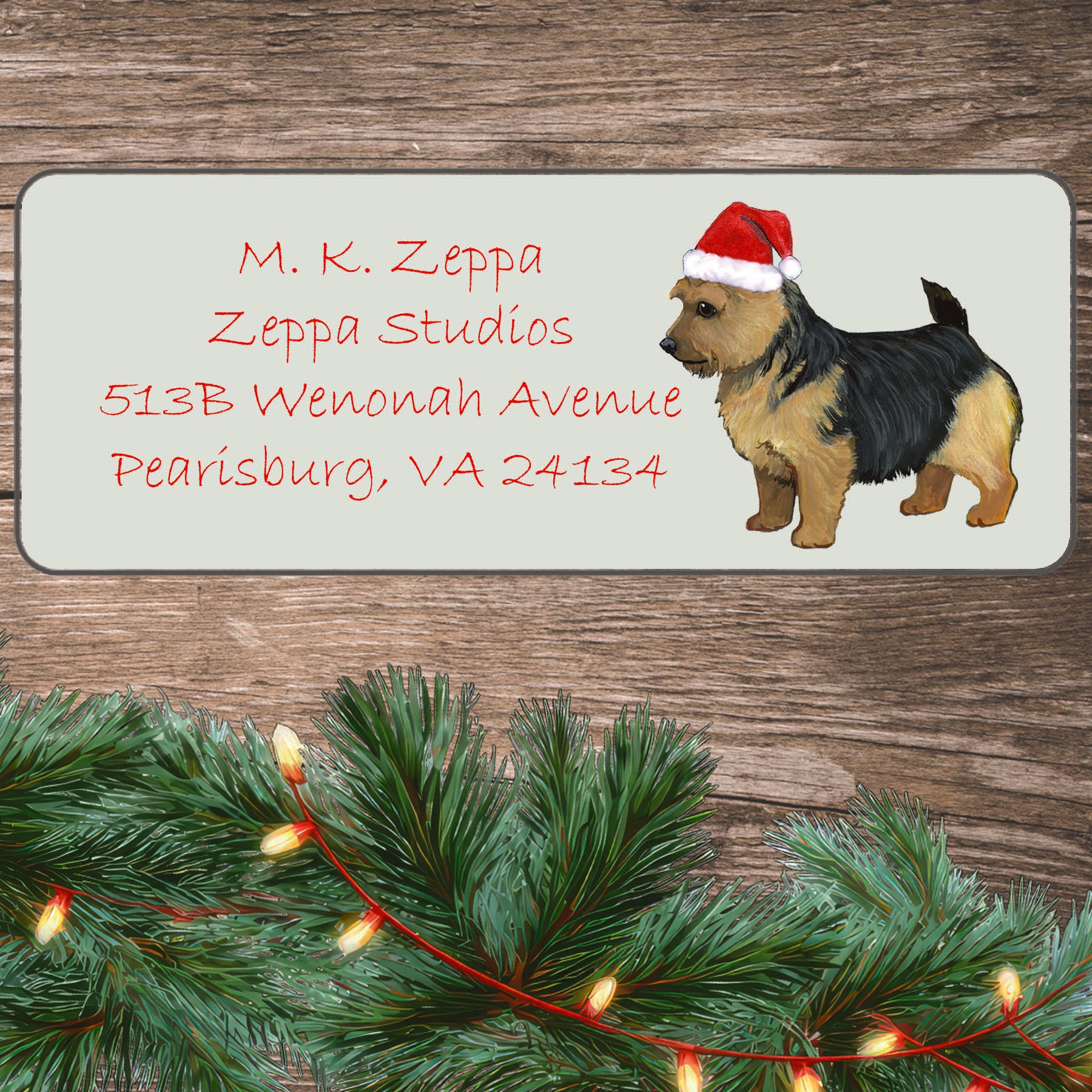 An illustration of a black and tan Norwich Terrier wearing a Santa Hat on an ivory return address label set off by a wood surface with holiday greenery background.