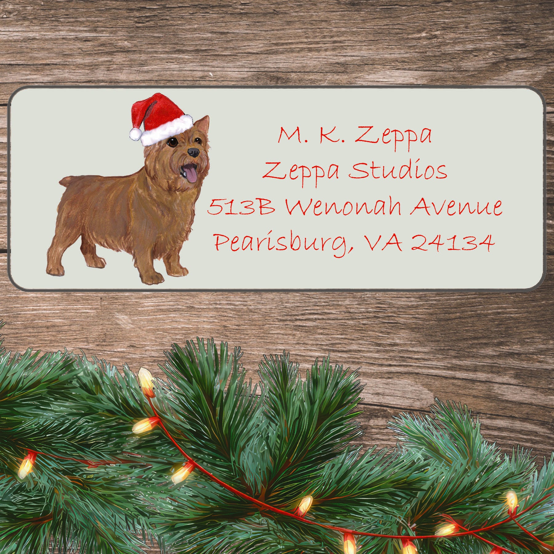 An illustration of a Norwich Terrier wearing a Santa Hat on an ivory return address label set off by a wood surface with holiday greenery background.