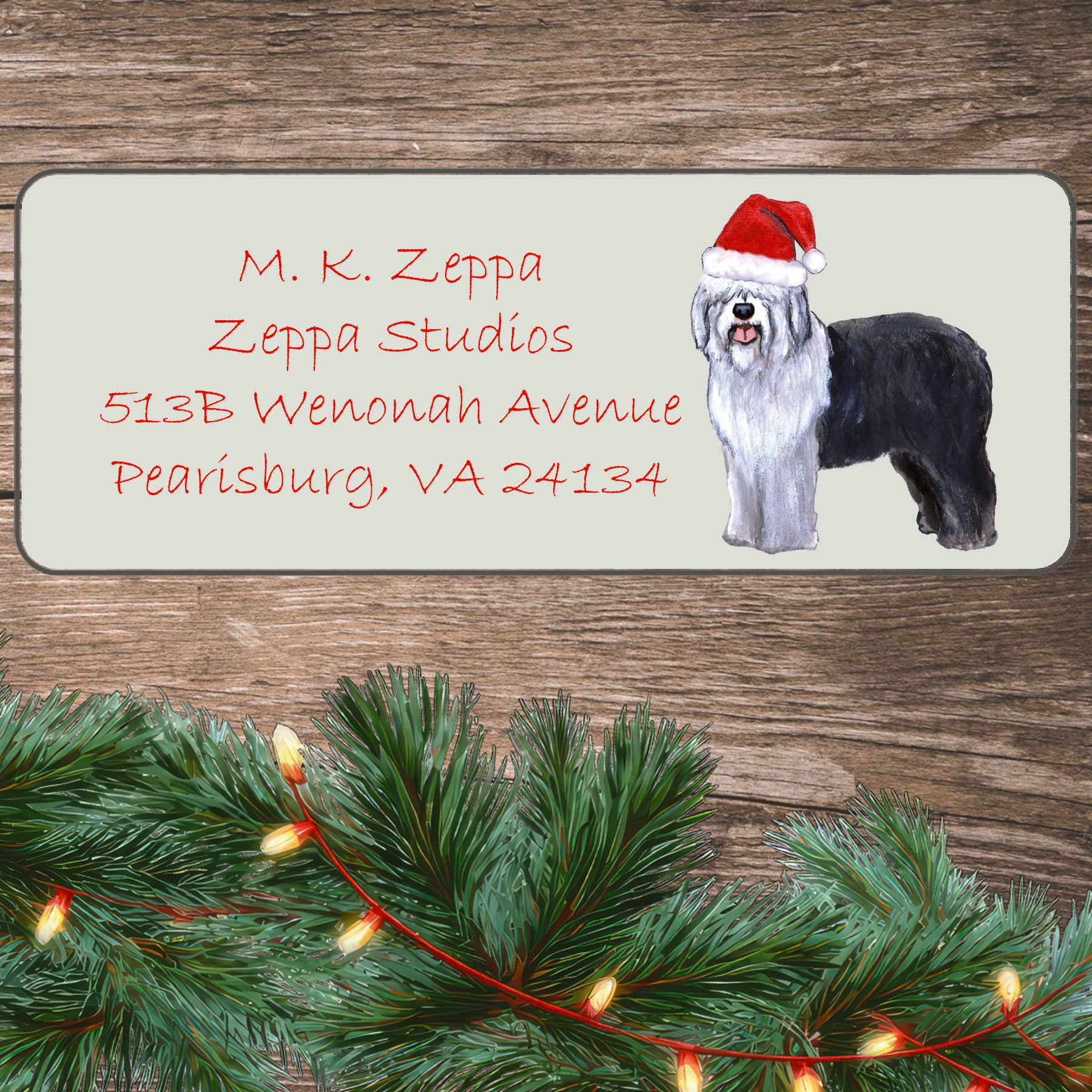 An illustration of an Old English Sheepdog wearing a Santa Hat on an ivory return address label set off by a wood surface with holiday greenery background.