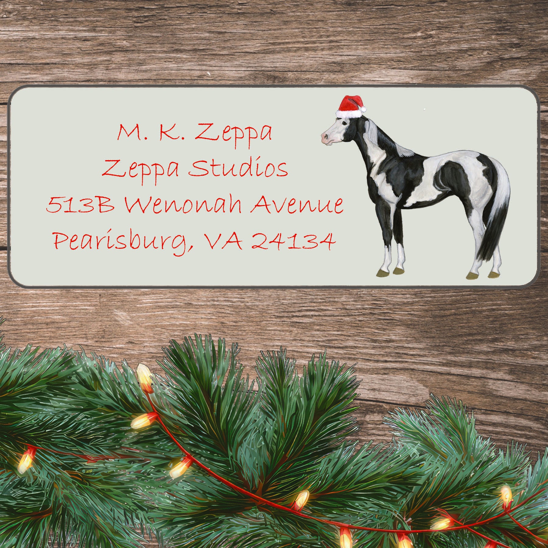 An illustration of a Paint Horse wearing a Santa Hat on an ivory return address label set off by a wood surface with holiday greenery background.