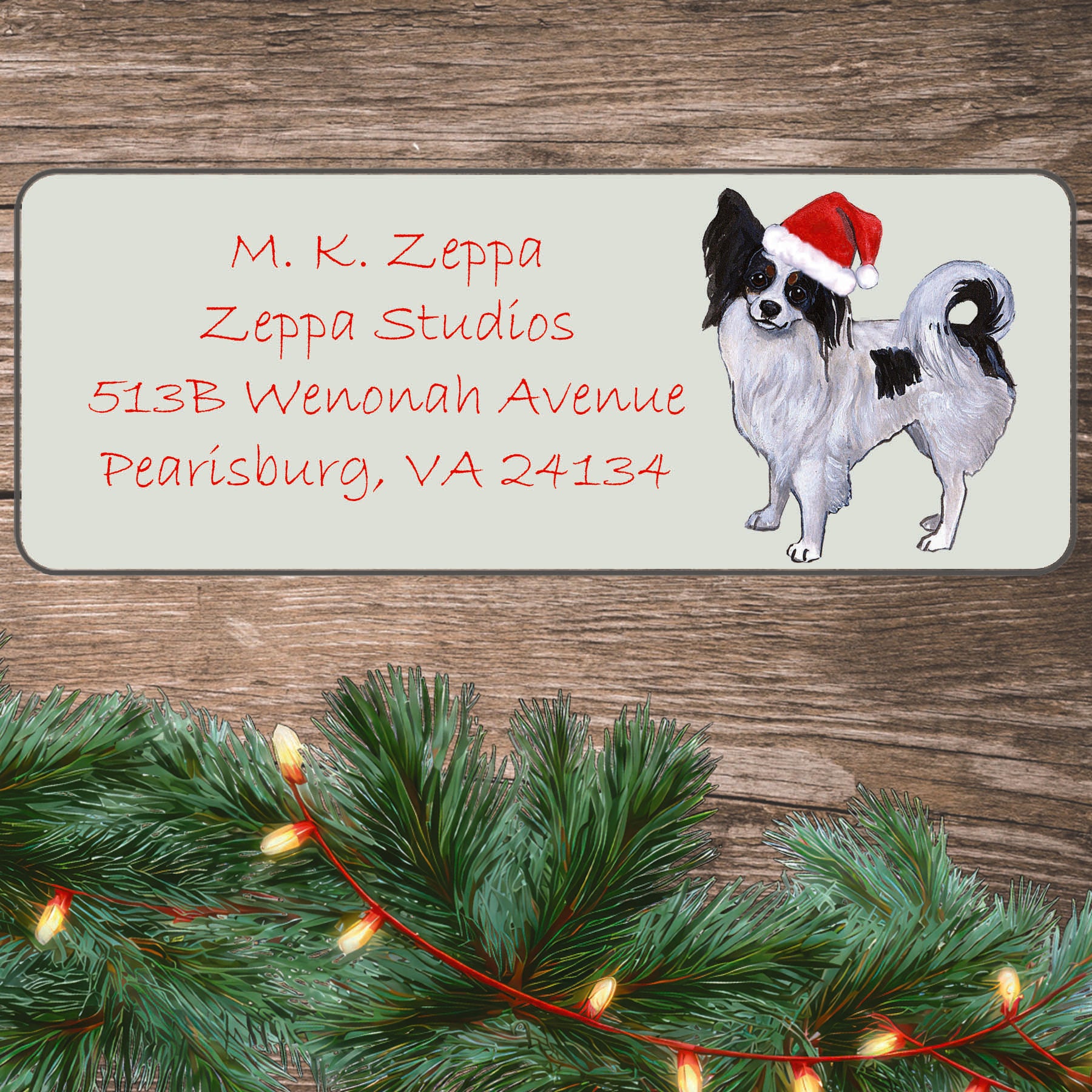 An illustration of a Papillon wearing a Santa Hat on an ivory return address label set off by a wood surface with holiday greenery background.