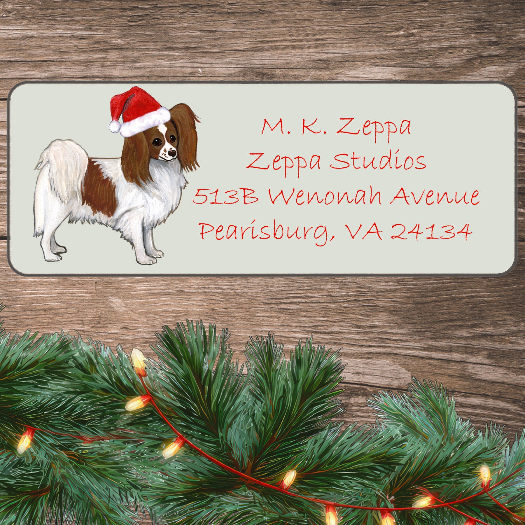 An illustration of a red and white Papillon wearing a Santa Hat on an ivory return address label set off by a wood surface with holiday greenery background.