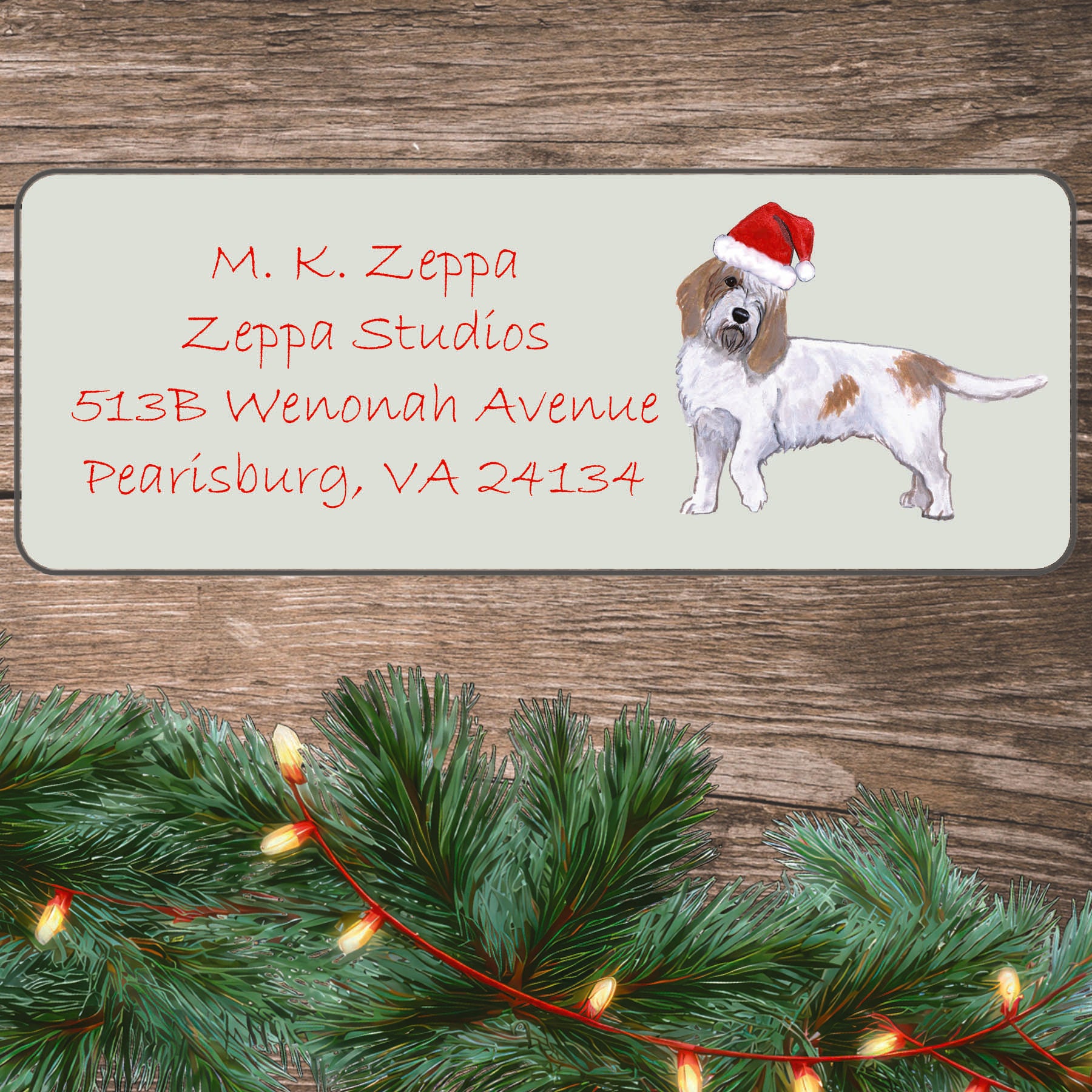 An illustration of a Petite Basset Griffon Vendeen wearing a Santa Hat on an ivory return address label set off by a wood surface with holiday greenery background.