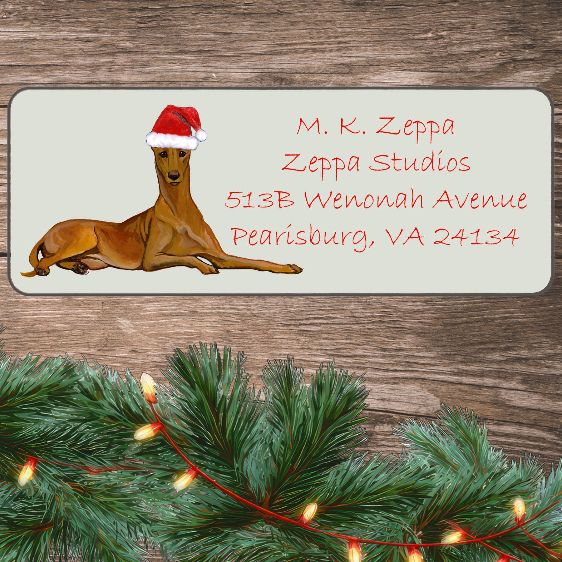 An illustration of a Pharaoh Hound wearing a Santa Hat on an ivory return address label set off by a wood surface with holiday greenery background.