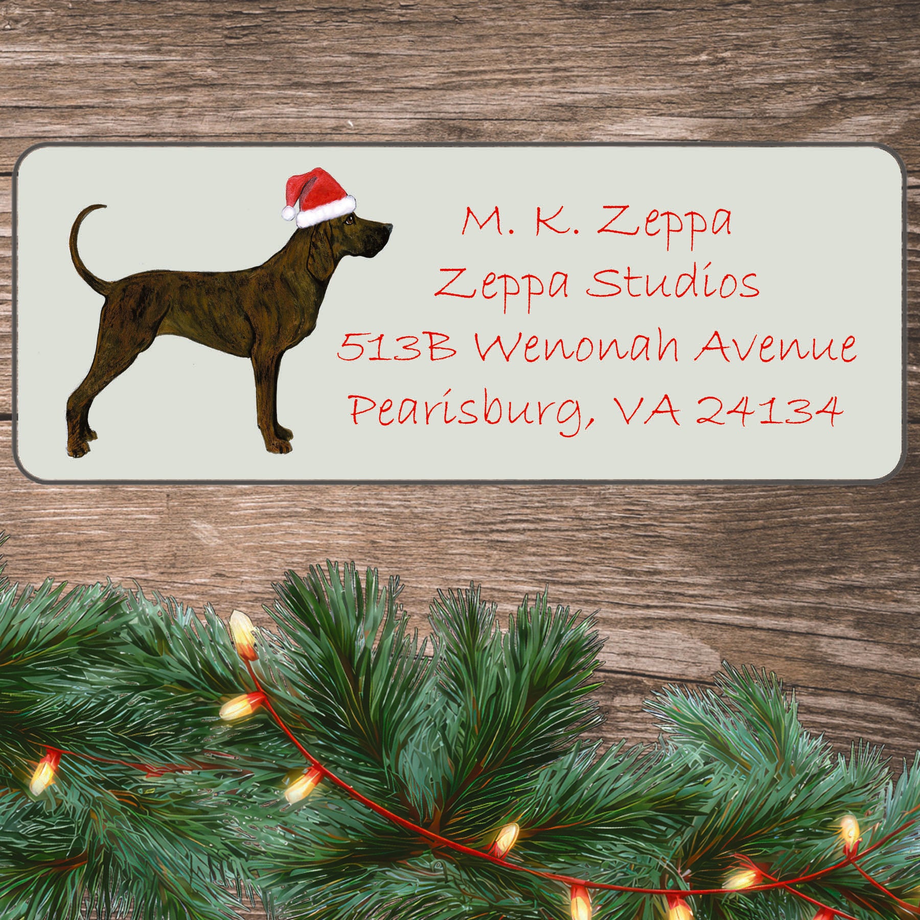 A Plott Hound in a Santa Hat illustration on an ivory return address label set off by a wood surface with holiday greenery background.
