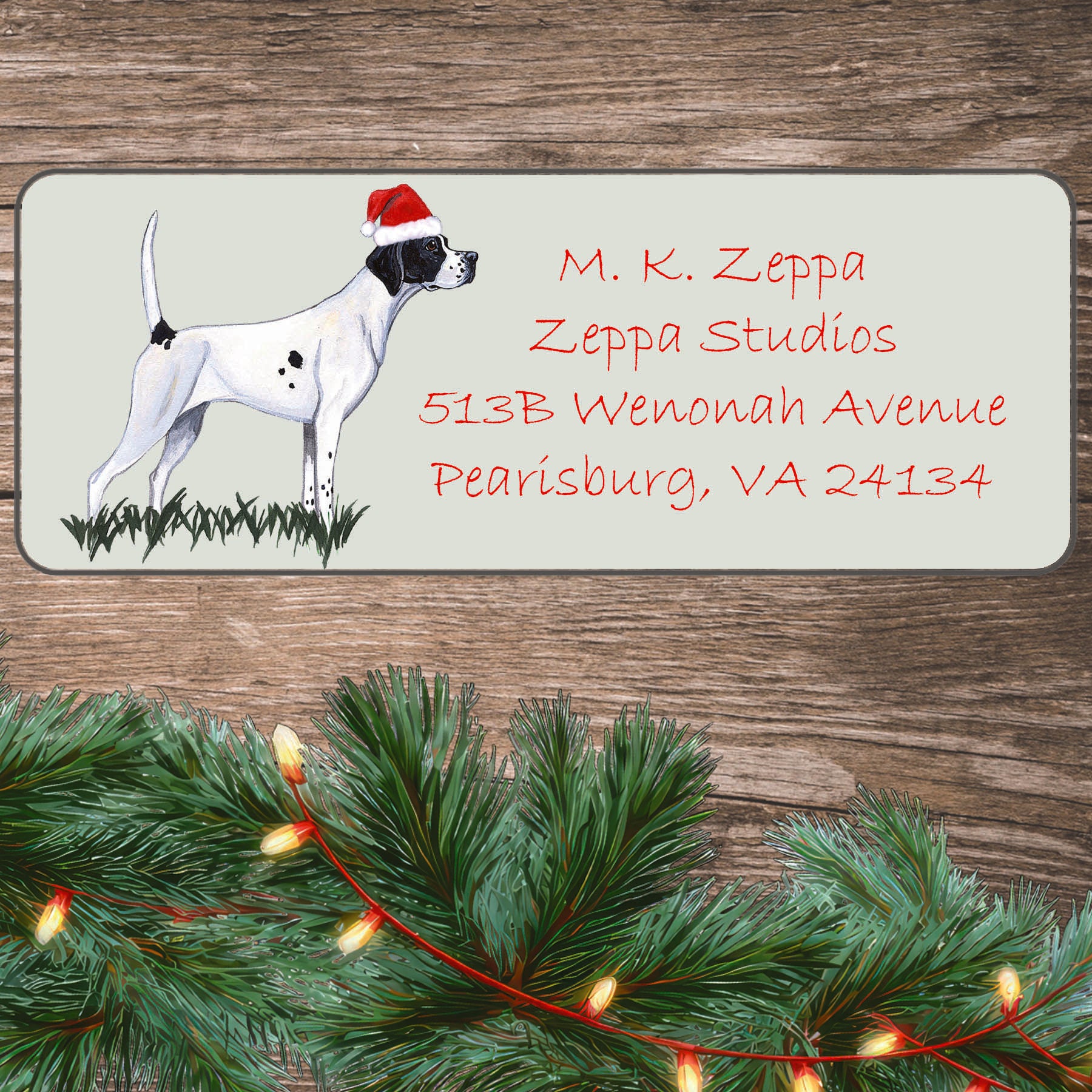 An illustration of a Pointer wearing a Santa Hat on an ivory return address label set off by a wood surface with holiday greenery background.