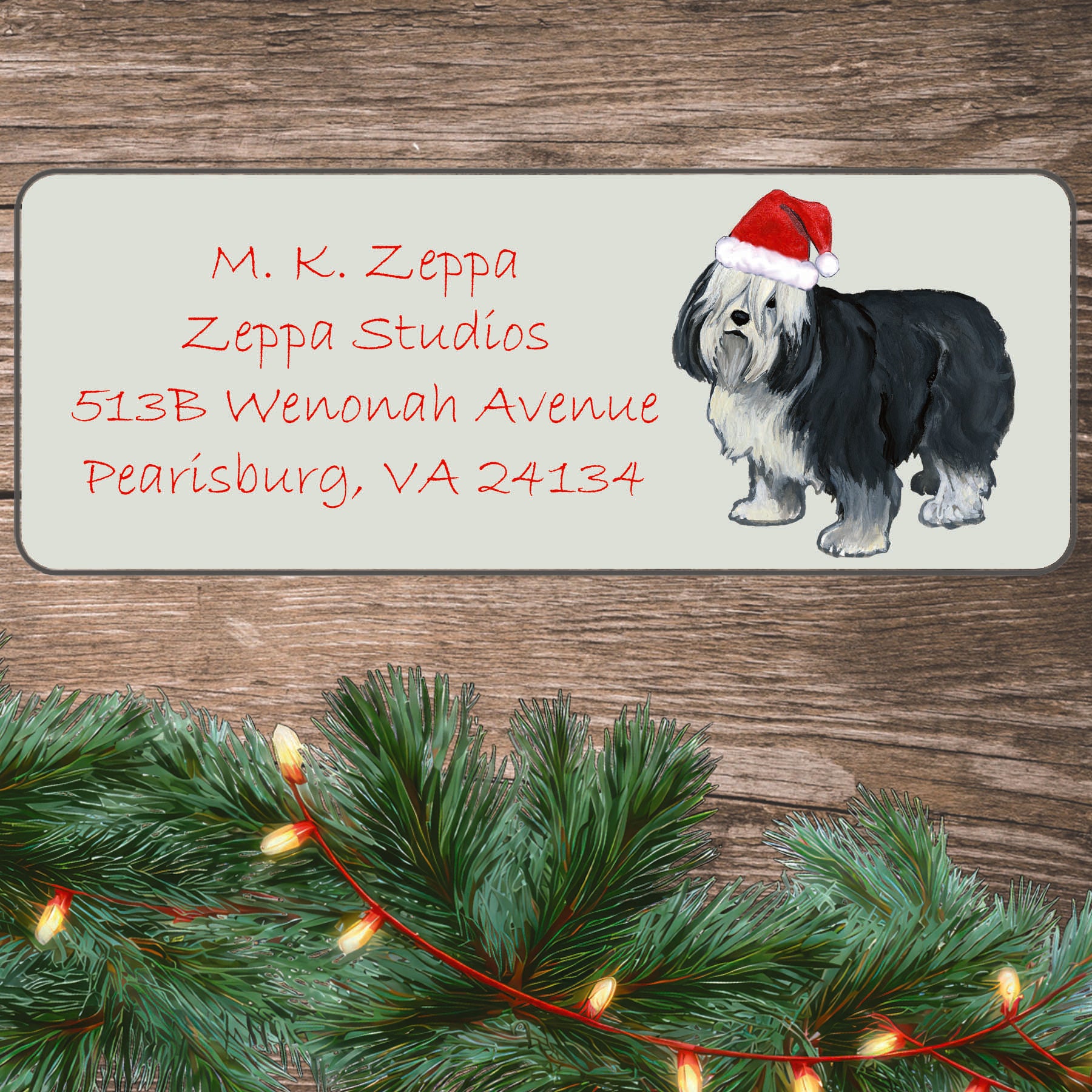 An illustration of a Polish Lowland Sheepdog wearing a Santa Hat on an ivory return address label set off by a wood surface with holiday greenery background.