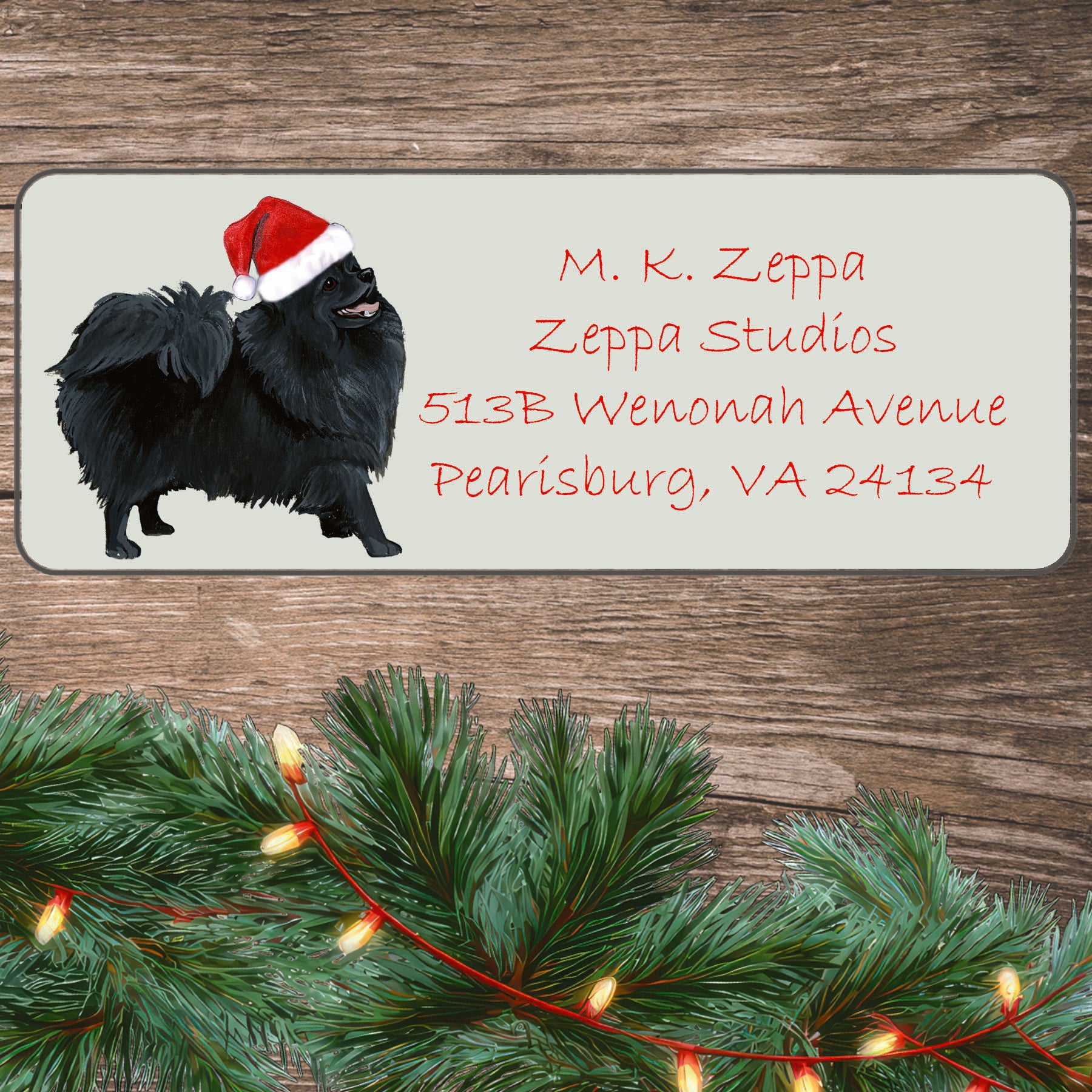 An illustration of a black Pomeranian wearing a Santa Hat on an ivory return address label set off by a wood surface with holiday greenery background.