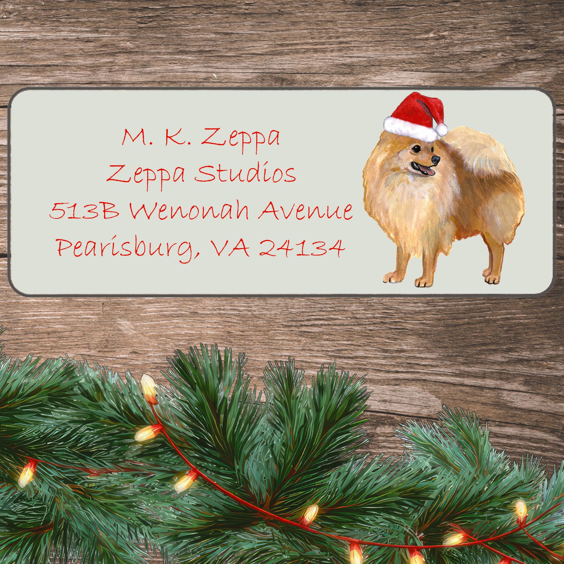 An illustration of a Pomeranian wearing a Santa Hat on an ivory return address label set off by a wood surface with holiday greenery background.