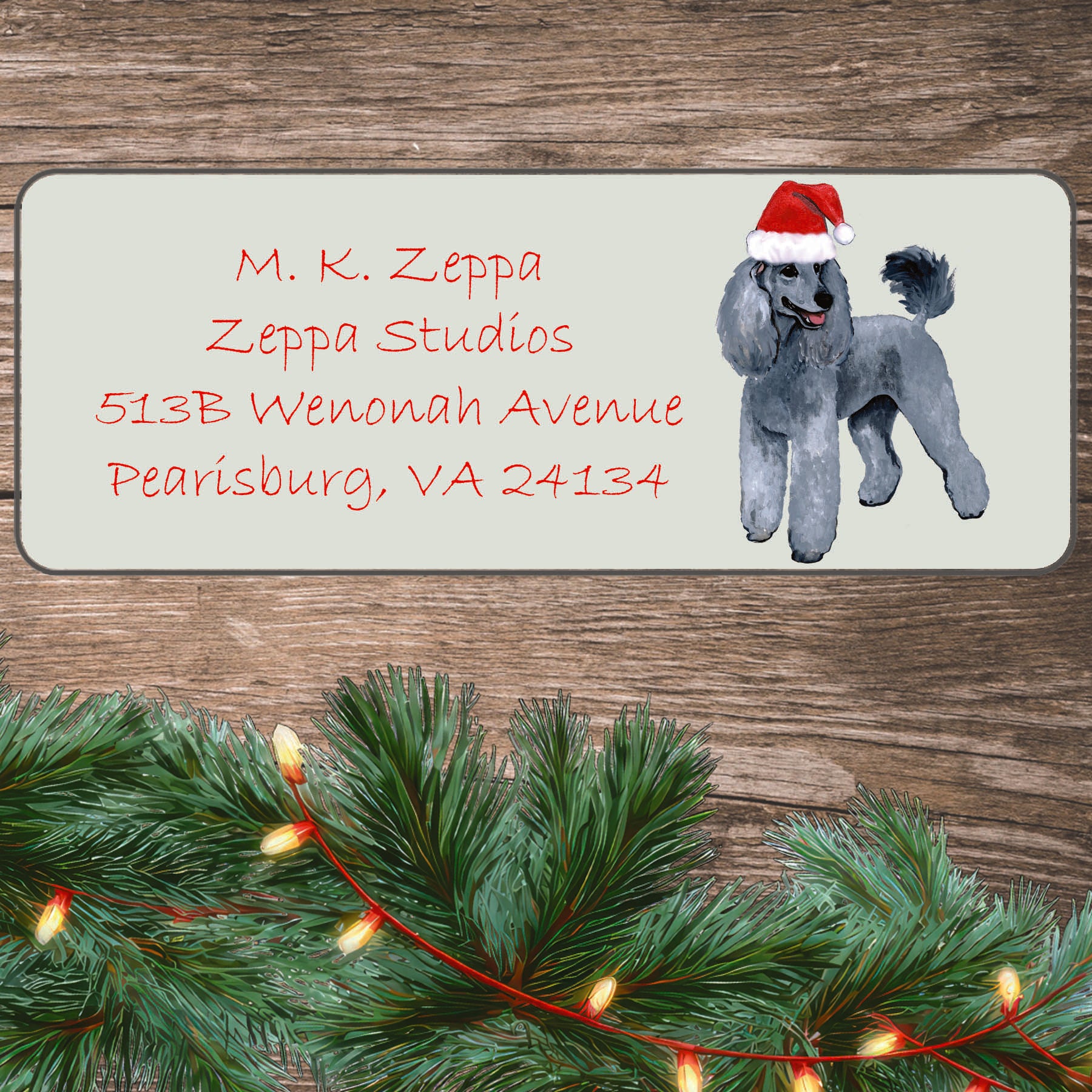 An illustration of a silver Poodle wearing a Santa Hat on an ivory return address label set off by a wood surface with holiday greenery background.