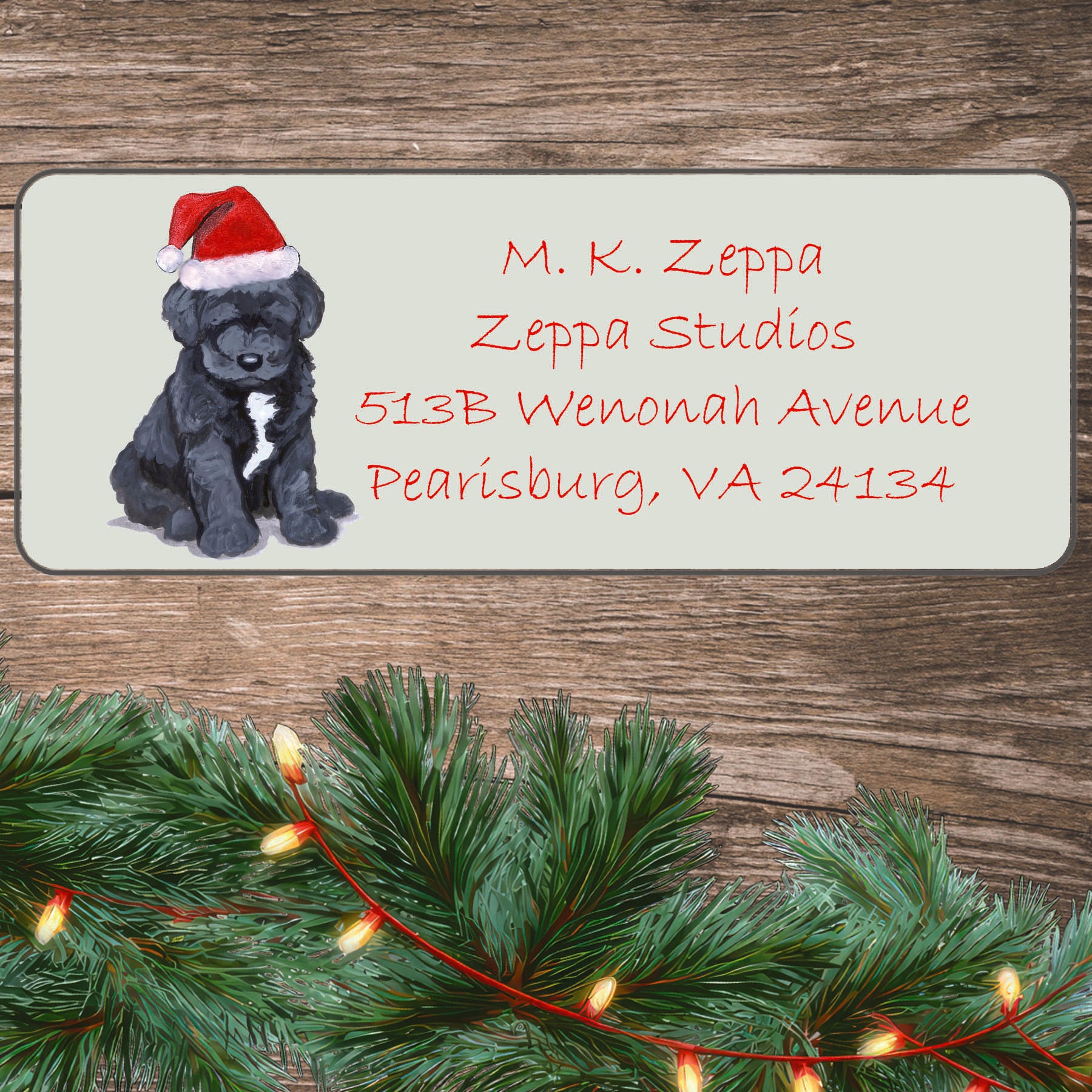 An illustration of a Portuguese Water Dog puppy wearing a Santa Hat on an ivory return address label set off by a wood surface with holiday greenery background.