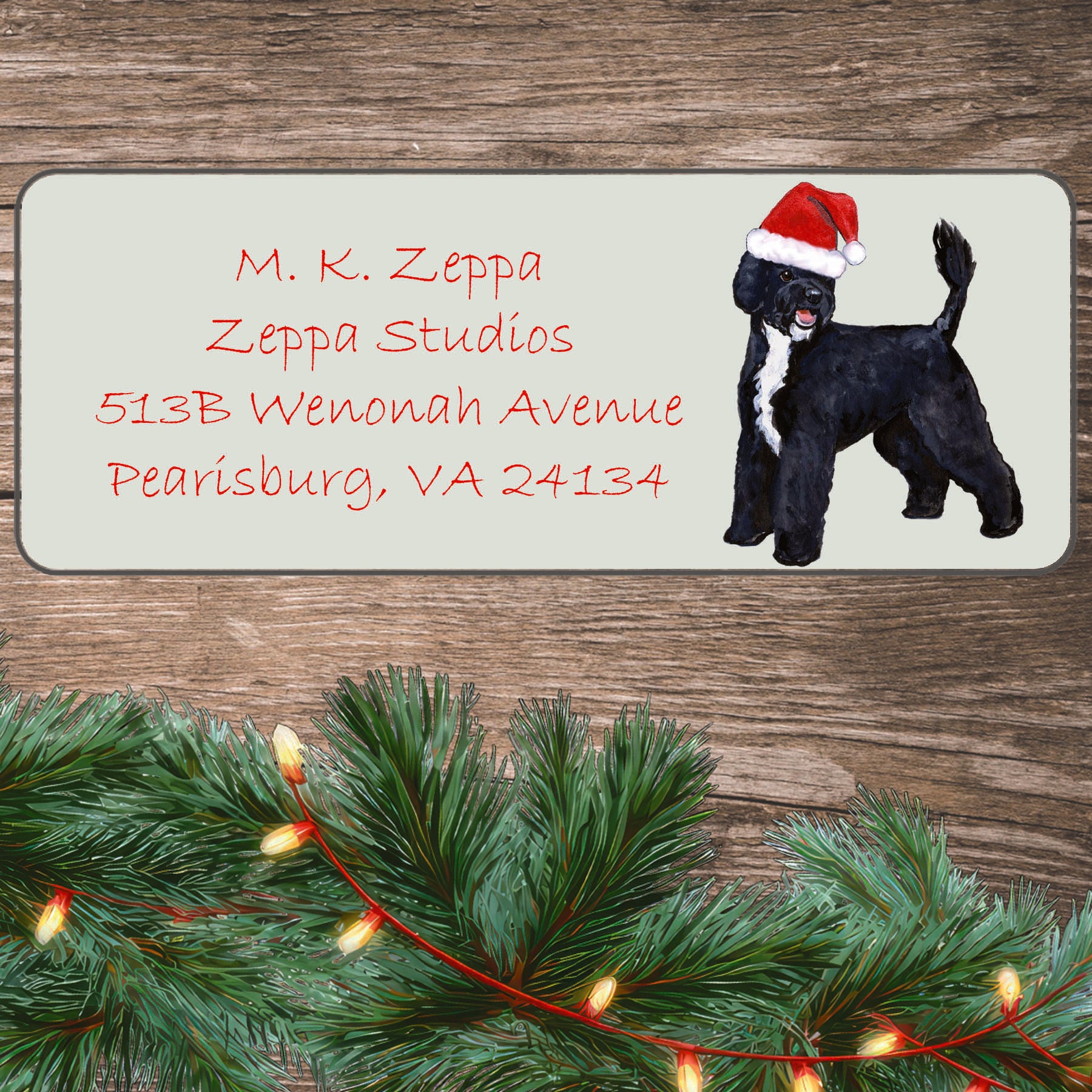 An illustration of a Portuguese Water Dog wearing a Santa Hat on an ivory return address label set off by a wood surface with holiday greenery background.