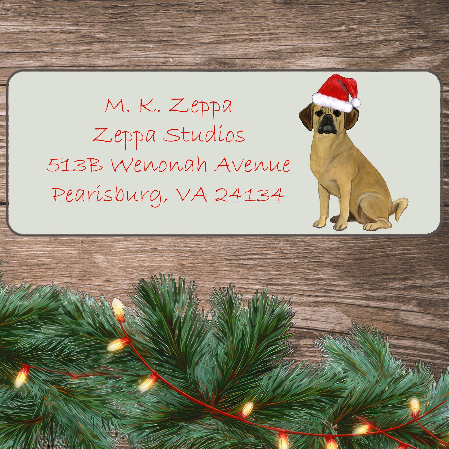 An illustration of a Puggle wearing a Santa Hat on an ivory return address label set off by a wood surface with holiday greenery background.