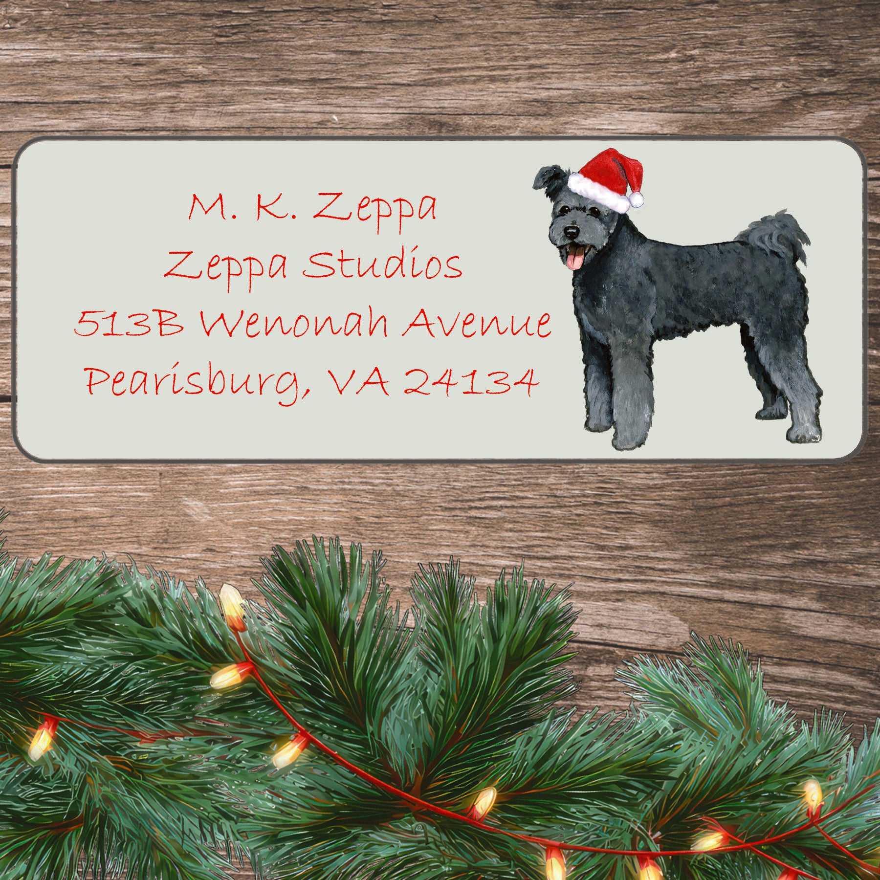 A Pumi in a Santa Hat illustration on an ivory return address label set off by a wood surface with holiday greenery background.