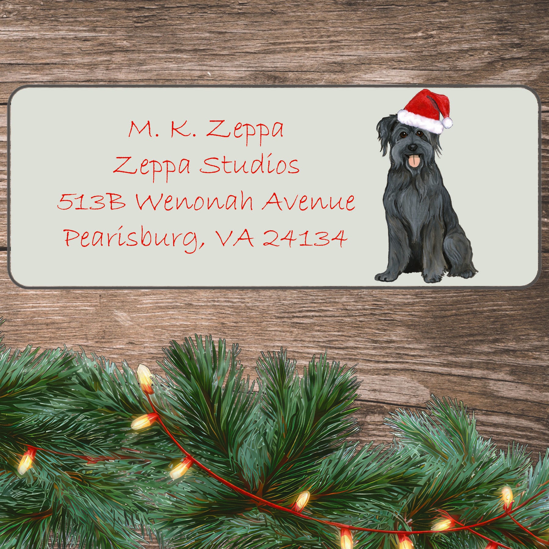An illustration of a Pyrenean Shepherd wearing a Santa Hat on an ivory return address label set off by a wood surface with holiday greenery background.