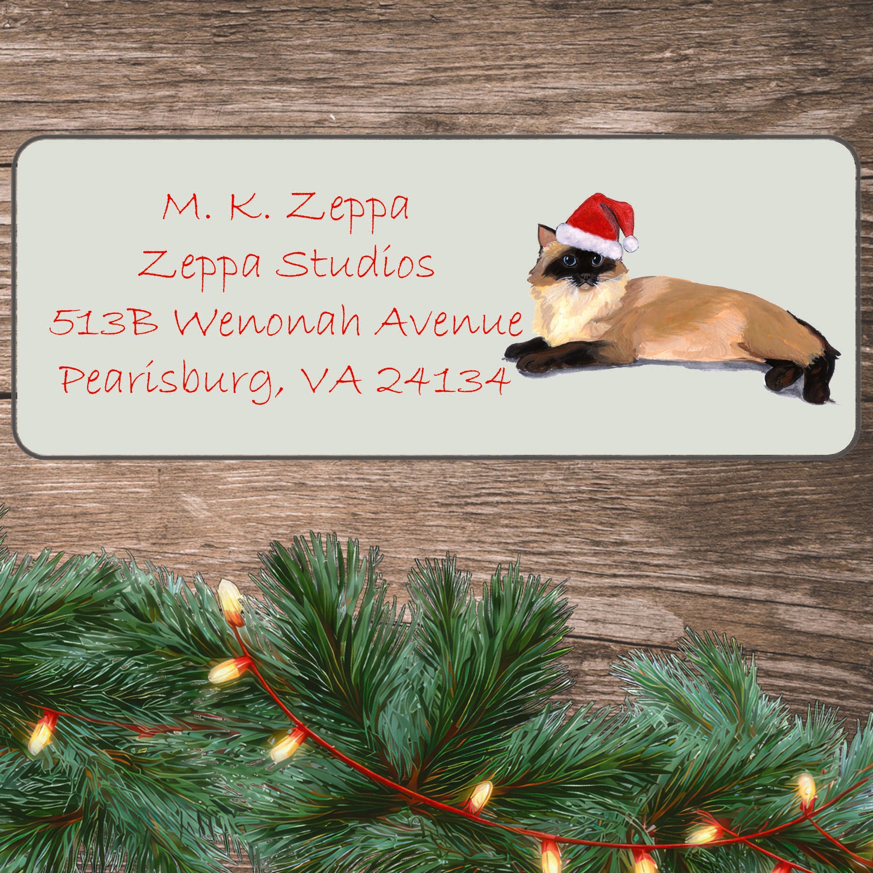 An illustration of a Ragdoll Cat  wearing a Santa Hat on an ivory return address label set off by a wood surface with holiday greenery background.