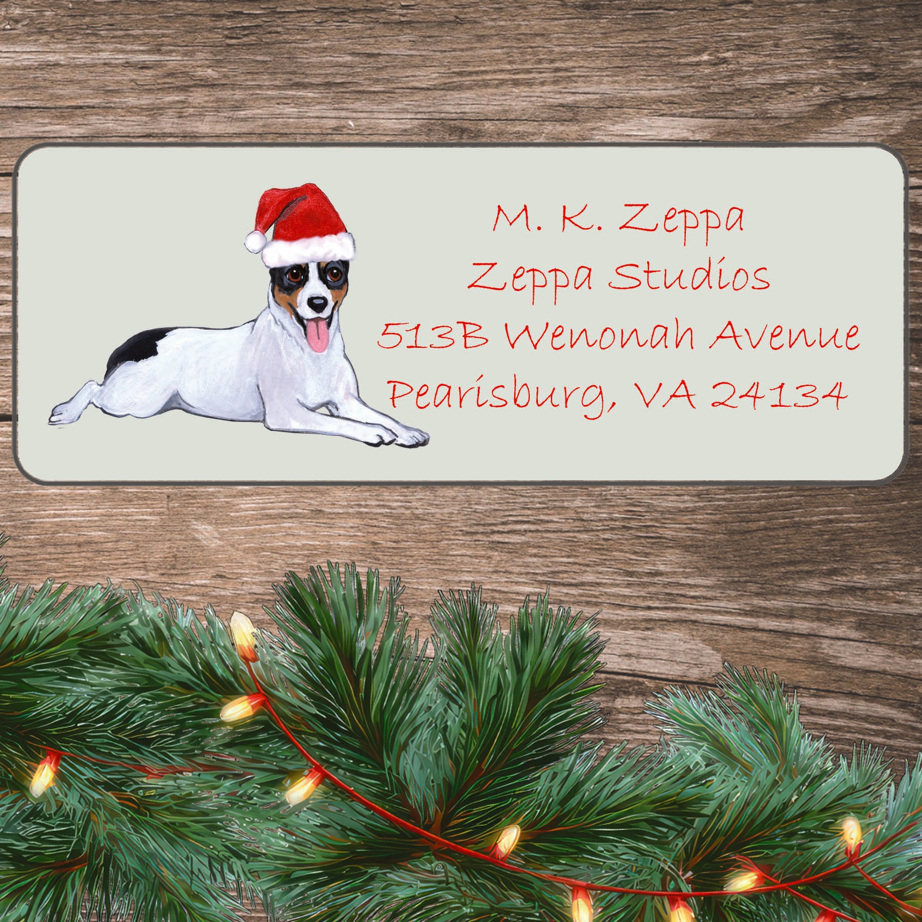 An illustration of a Rat Terrier wearing a Santa Hat on an ivory return address label set off by a wood surface with holiday greenery background.
