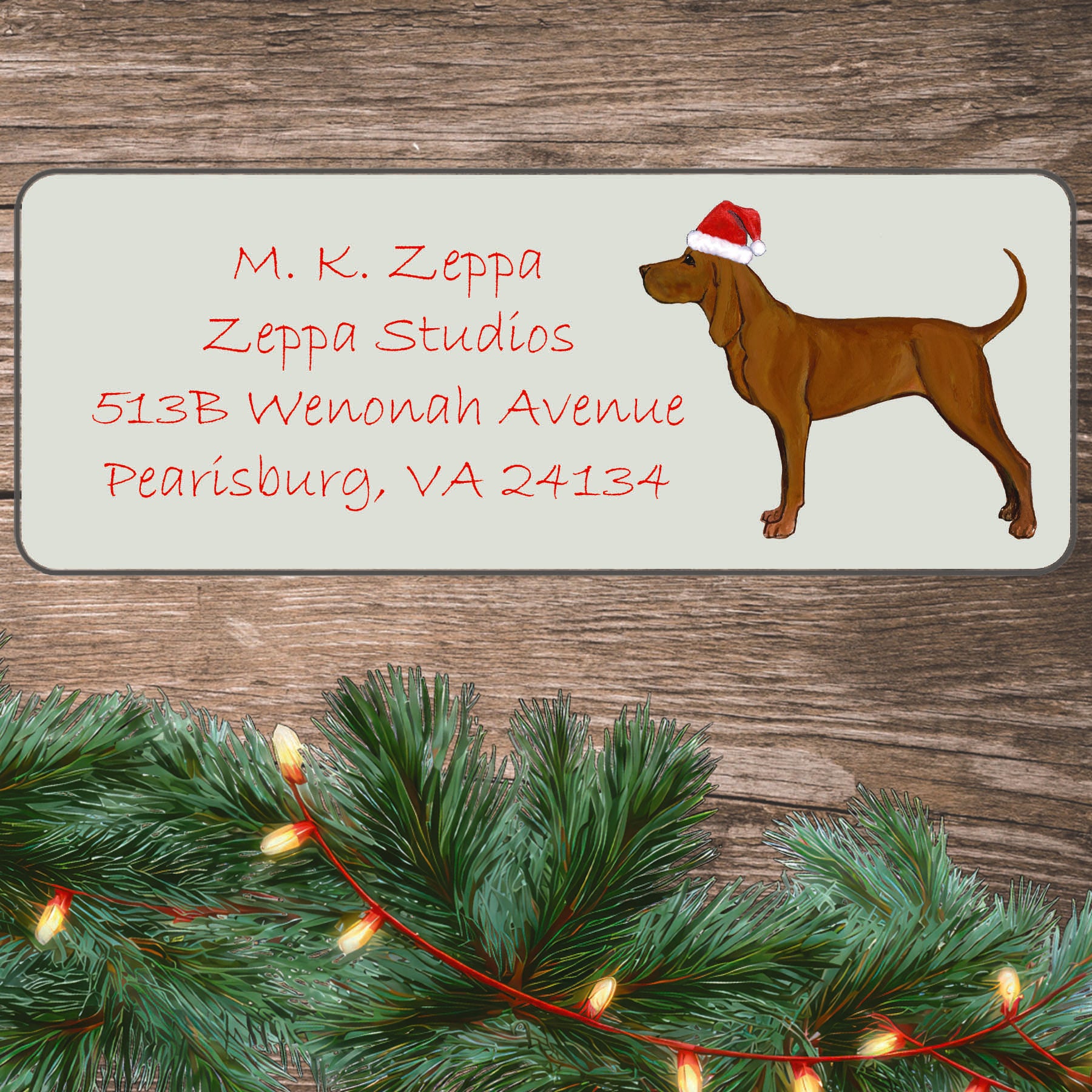 An illustration of a Redbone Coonhound wearing a Santa Hat on an ivory return address label set off by a wood surface with holiday greenery background.