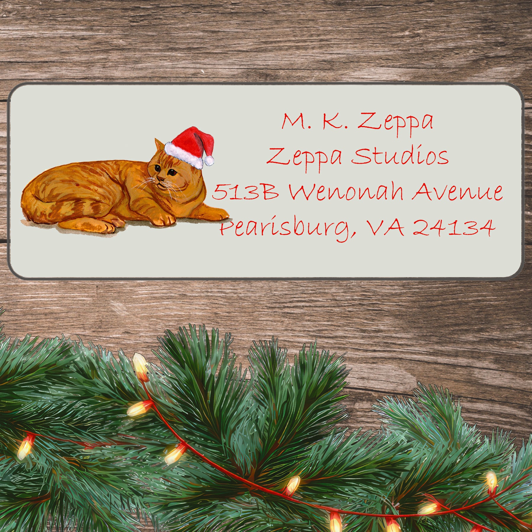 An illustration of an Orange Tabby Cat wearing a Santa Hat on an ivory return address label set off by a wood surface with holiday greenery background.