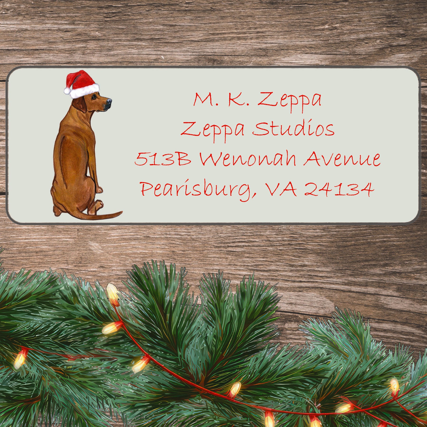 An illustration of a Rhodesian Ridgeback wearing a Santa Hat on an ivory return address label set off by a wood surface with holiday greenery background.