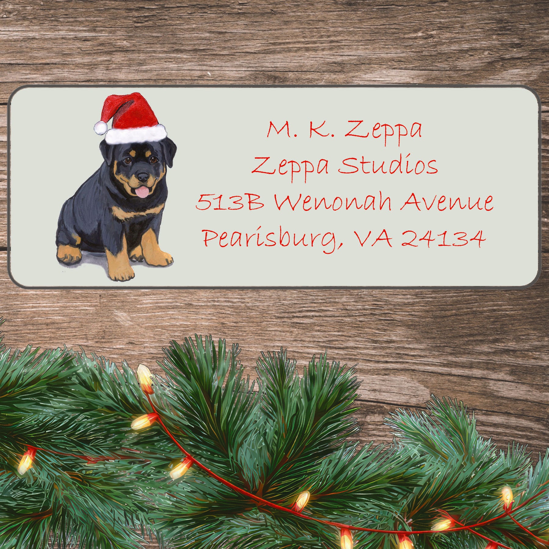 An illustration of a Rottweiler Puppy wearing a Santa Hat on an ivory return address label set off by a wood surface with holiday greenery background.