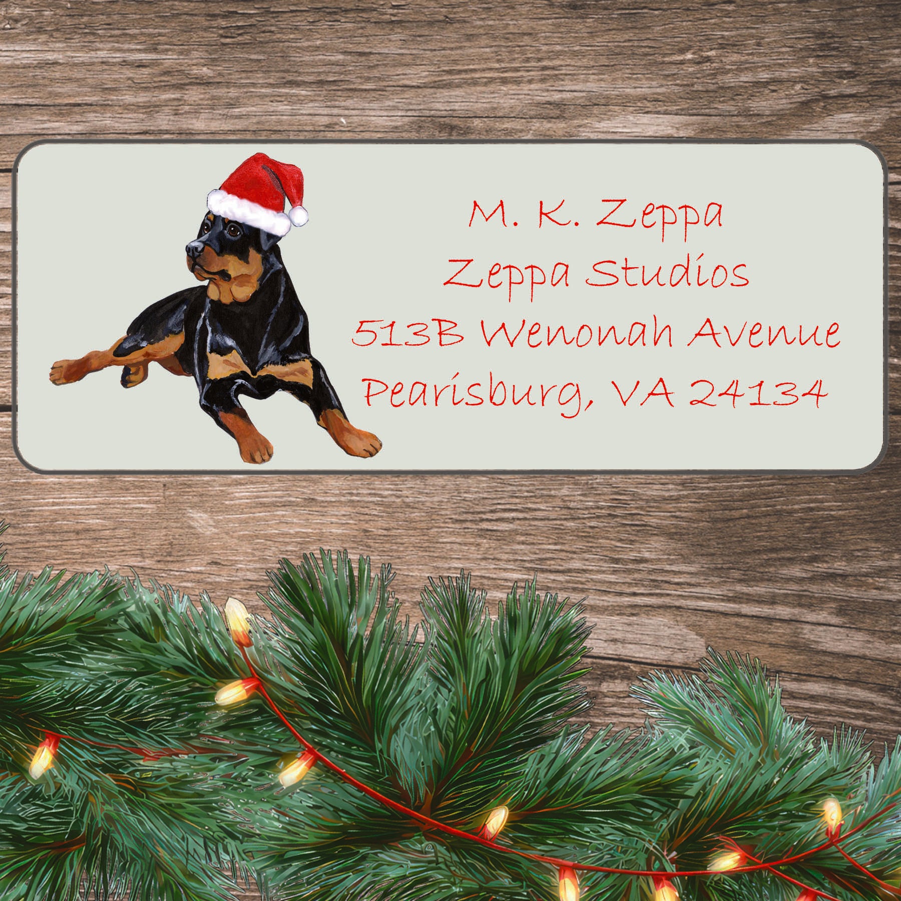 An illustration of a Rottweiler wearing a Santa Hat on an ivory return address label set off by a wood surface with holiday greenery background.