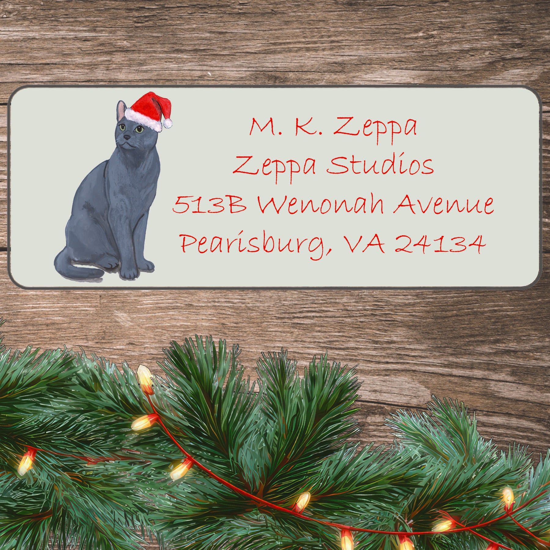 An illustration of a Russian Blue Cat wearing a Santa Hat on an ivory return address label set off by a wood surface with holiday greenery background.