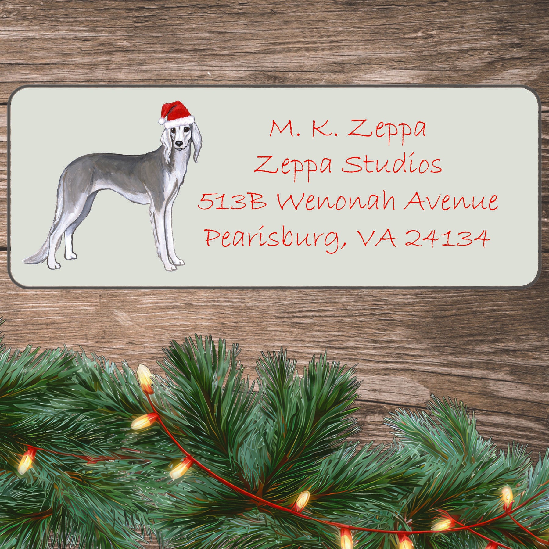 An illustration of a Saluki wearing a Santa Hat on an ivory return address label set off by a wood surface with holiday greenery background.
