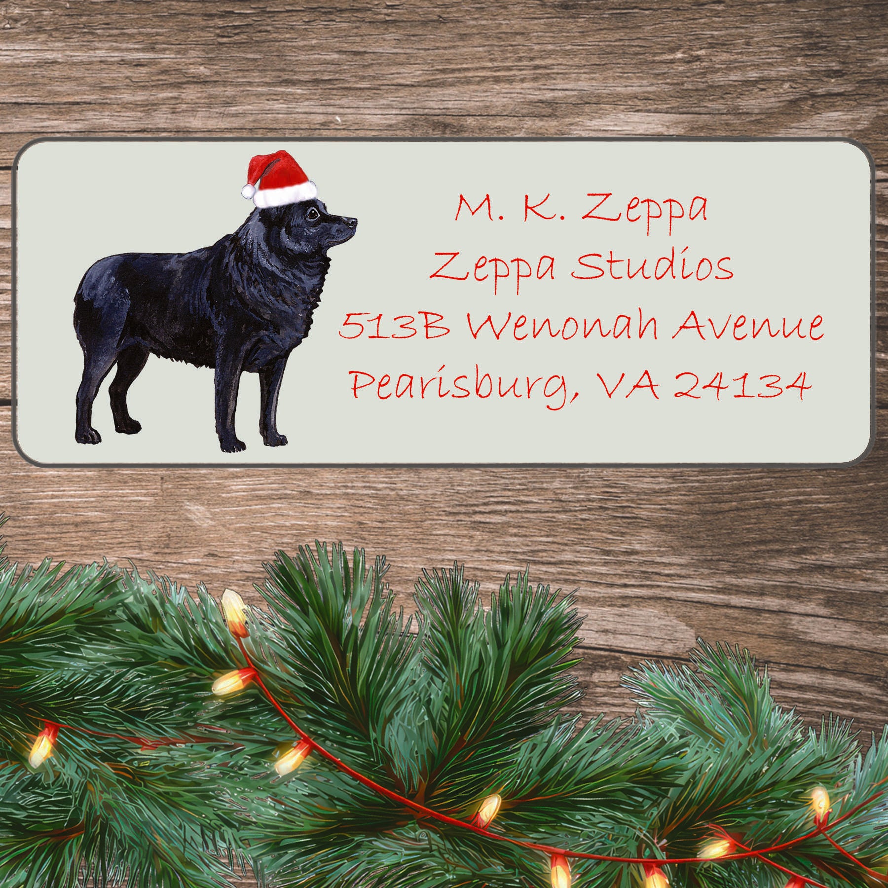 An illustration of a Schipperke wearing a Santa Hat on an ivory return address label set off by a wood surface with holiday greenery background.