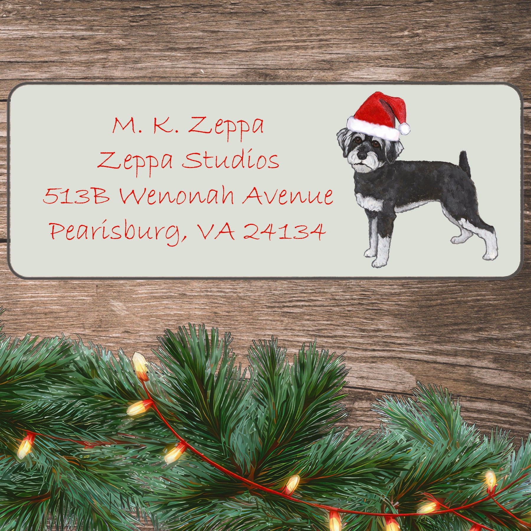 A Schnoodle in a Santa Hat illustration on an ivory return address label set off by a wood surface with holiday greenery background.