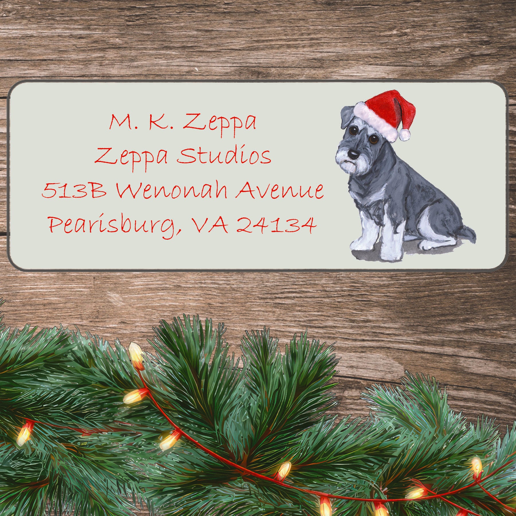 An illustration of a Schnauzer puppy wearing a Santa Hat on an ivory return address label set off by a wood surface with holiday greenery background.