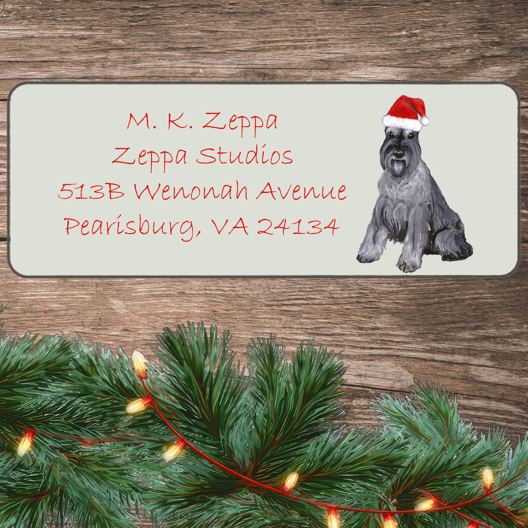 An illustration of a Schnauzer wearing a Santa Hat on an ivory return address label set off by a wood surface with holiday greenery background.