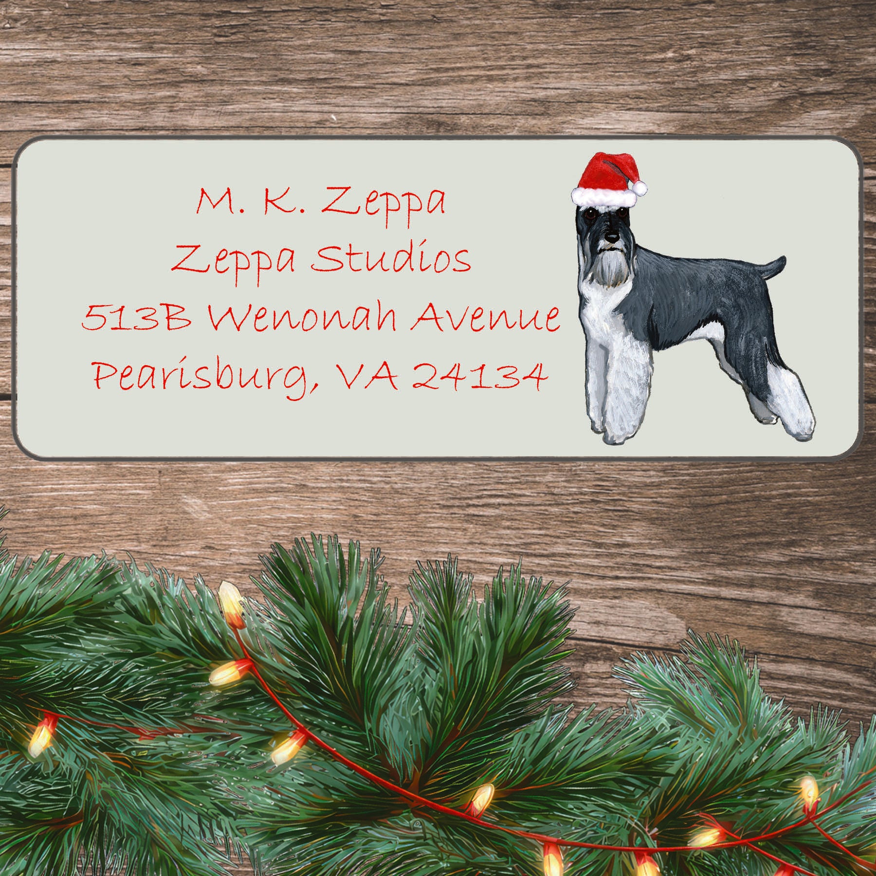 An illustration of a standing Schnauzer wearing a Santa Hat on an ivory return address label set off by a wood surface with holiday greenery background.