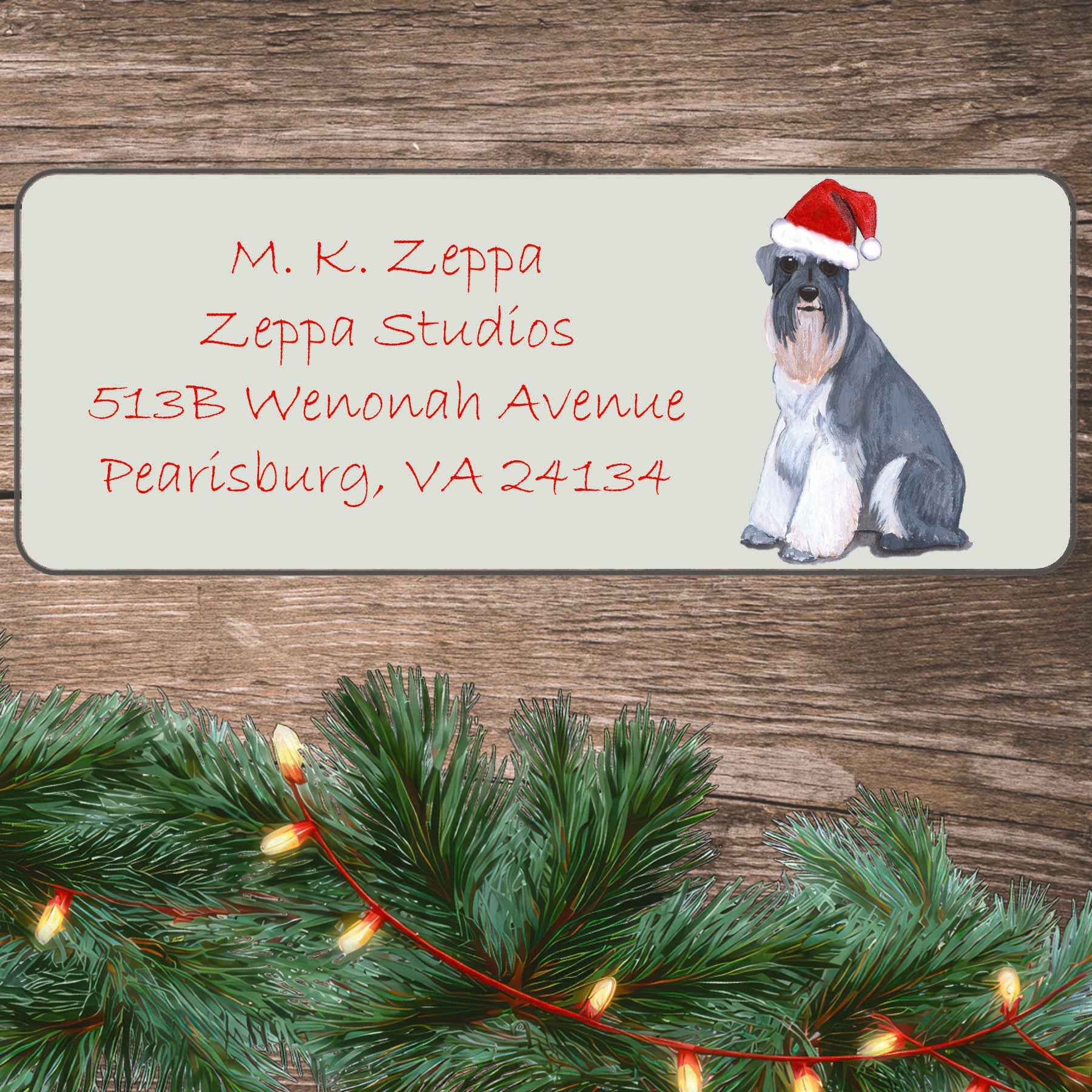 An illustration of a Schnauzer wearing a Santa Hat on an ivory return address label set off by a wood surface with holiday greenery background.