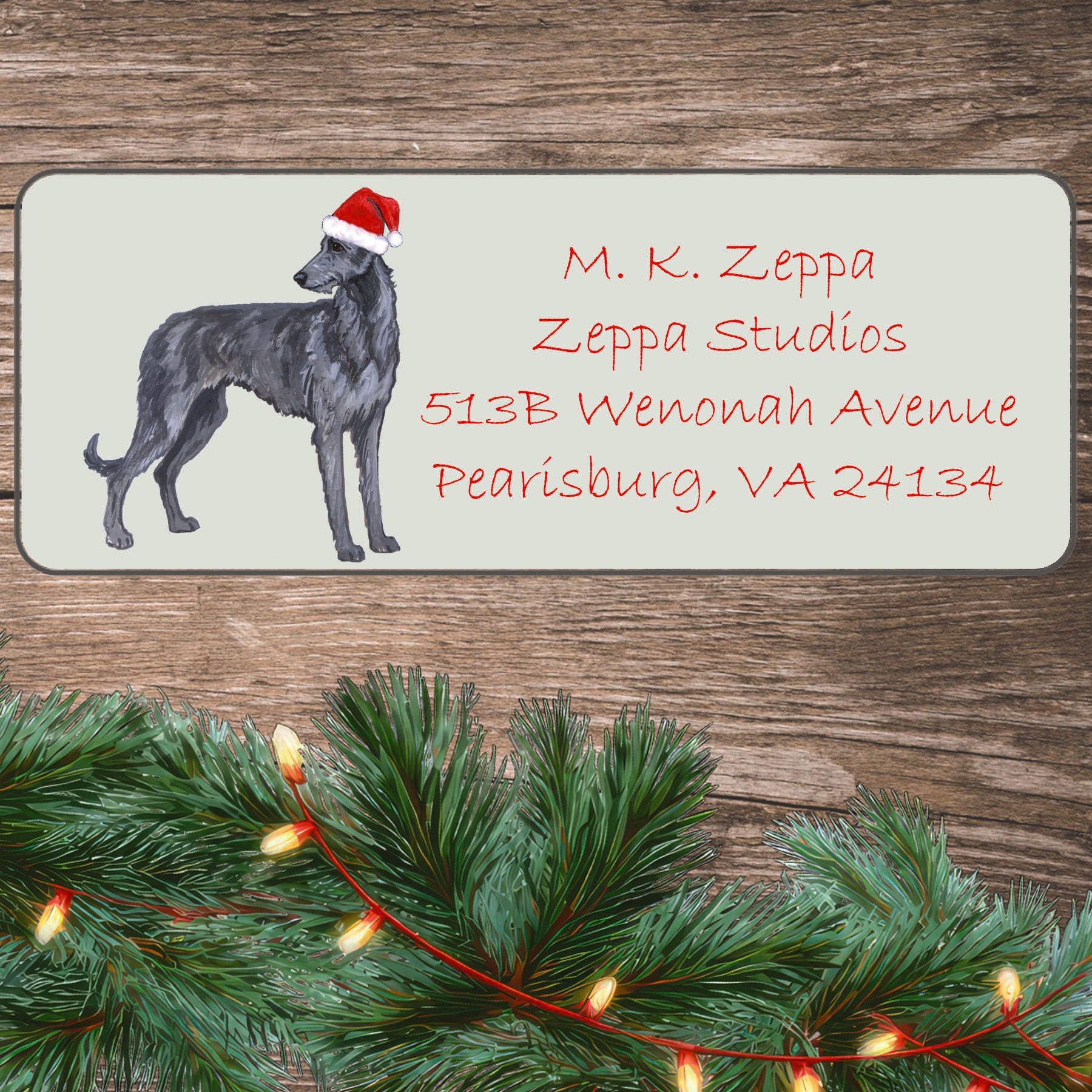 An illustration of a Scottish Deerhound wearing a Santa Hat on an ivory return address label set off by a wood surface with holiday greenery background.