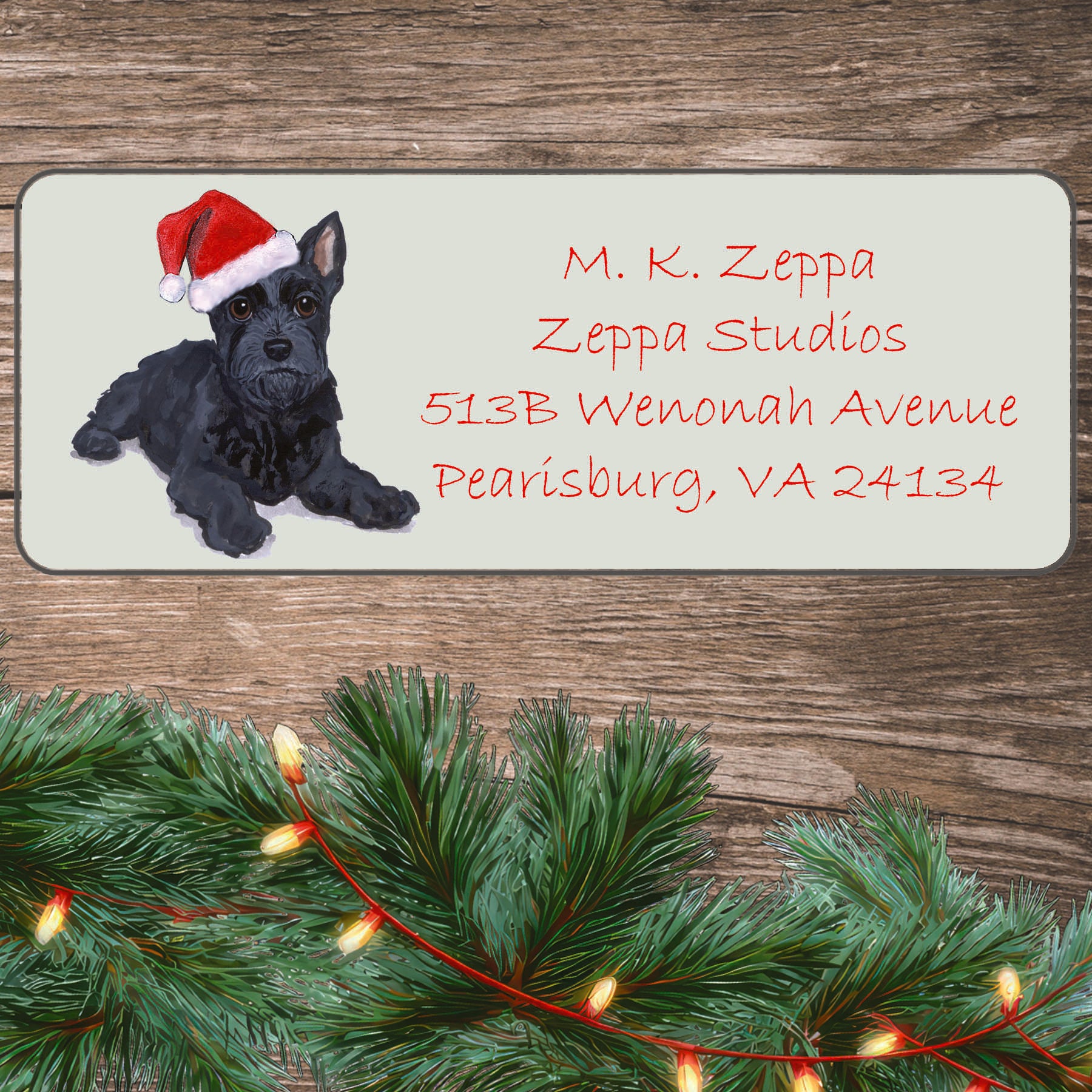 An illustration of a Scottish Terrier Puppy wearing a Santa Hat on an ivory return address label set off by a wood surface with holiday greenery background.